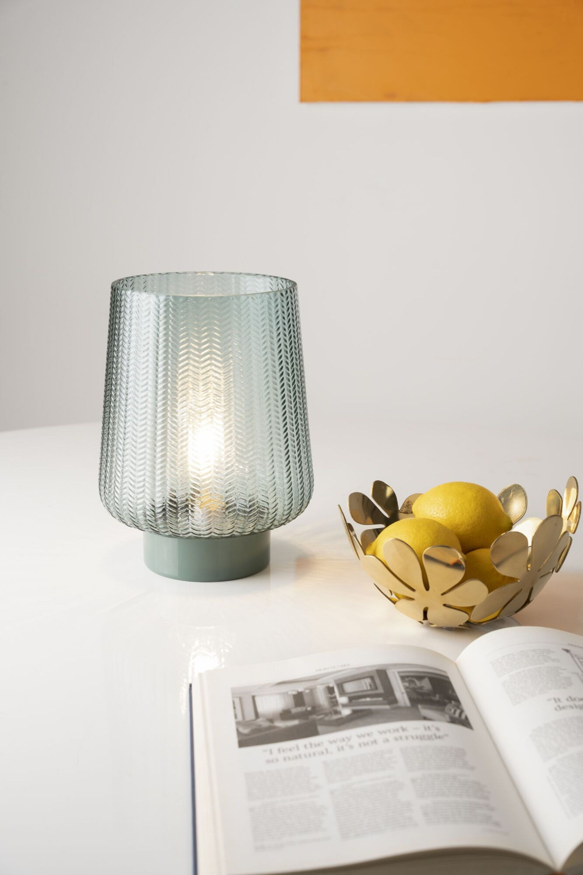 Niebiesko-zielona szklana lampa z białym światłem, złota miska z cytrynami i otwarta książka.