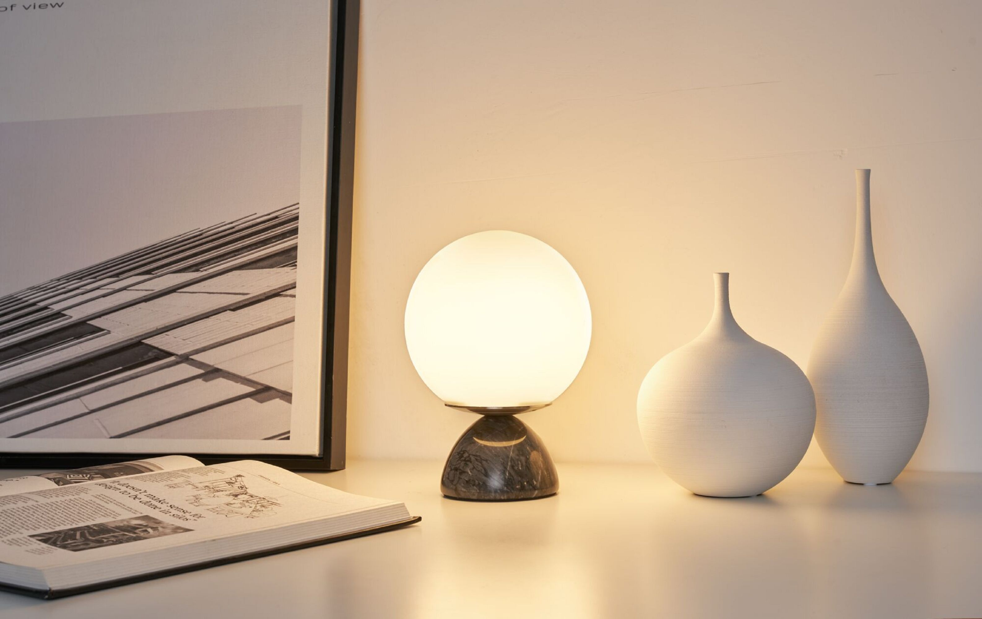 Eine Lampe mit Marmorsockel und weißem Globus-Schirm auf weißer Oberfläche. Bücher und Vasen sind im Hintergrund.