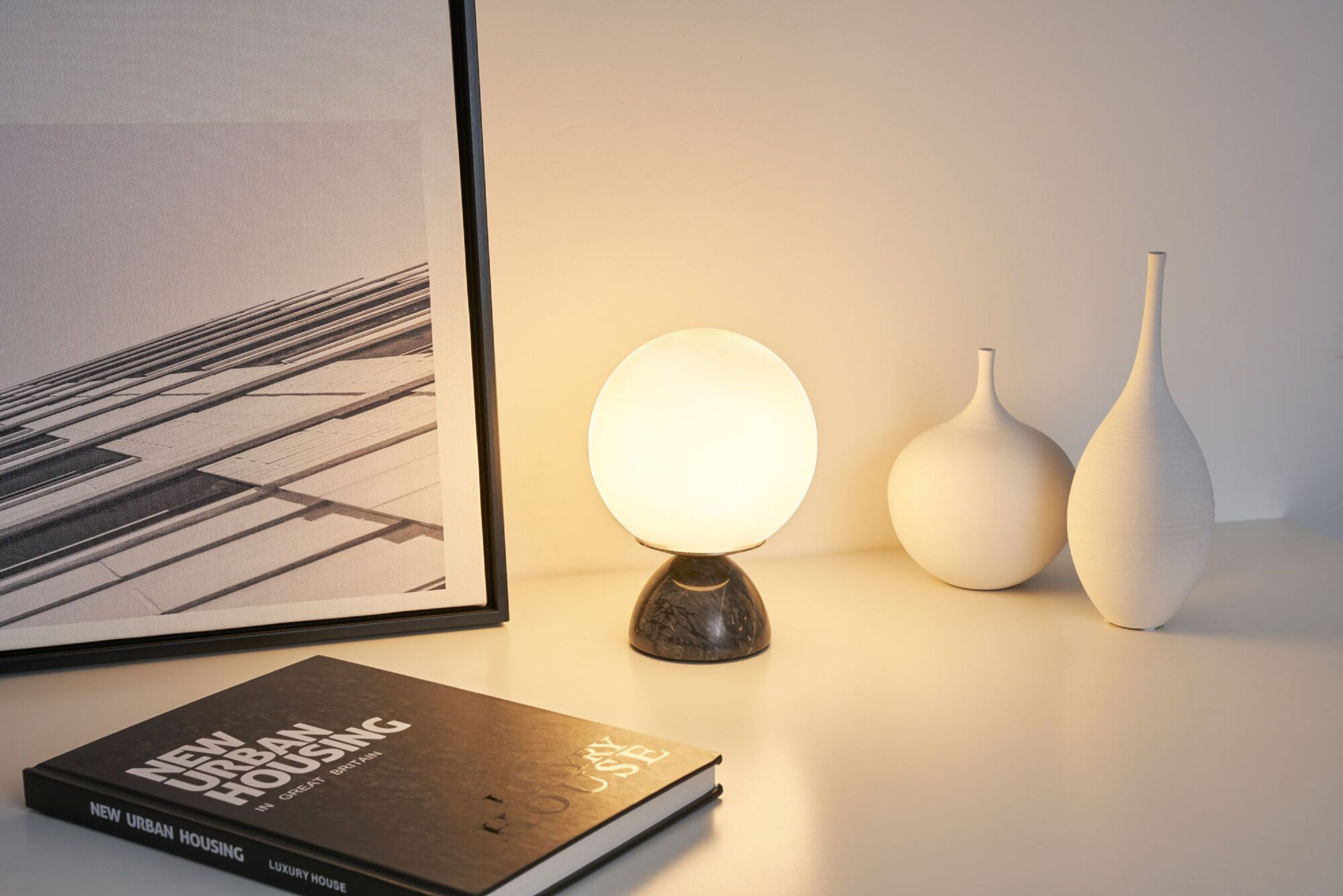 Eine beleuchtete Tischlampe mit Marmorsockel, ein Buch und Vasen auf weißer Fläche.