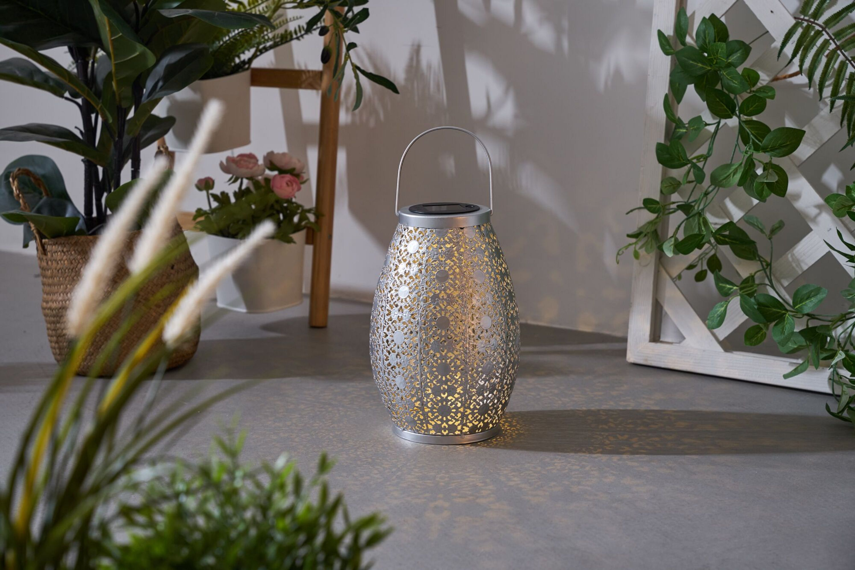 Srebrna latarnia z kwiatowym wzorem, otoczona roślinami. Szara podłoga, białe tło.