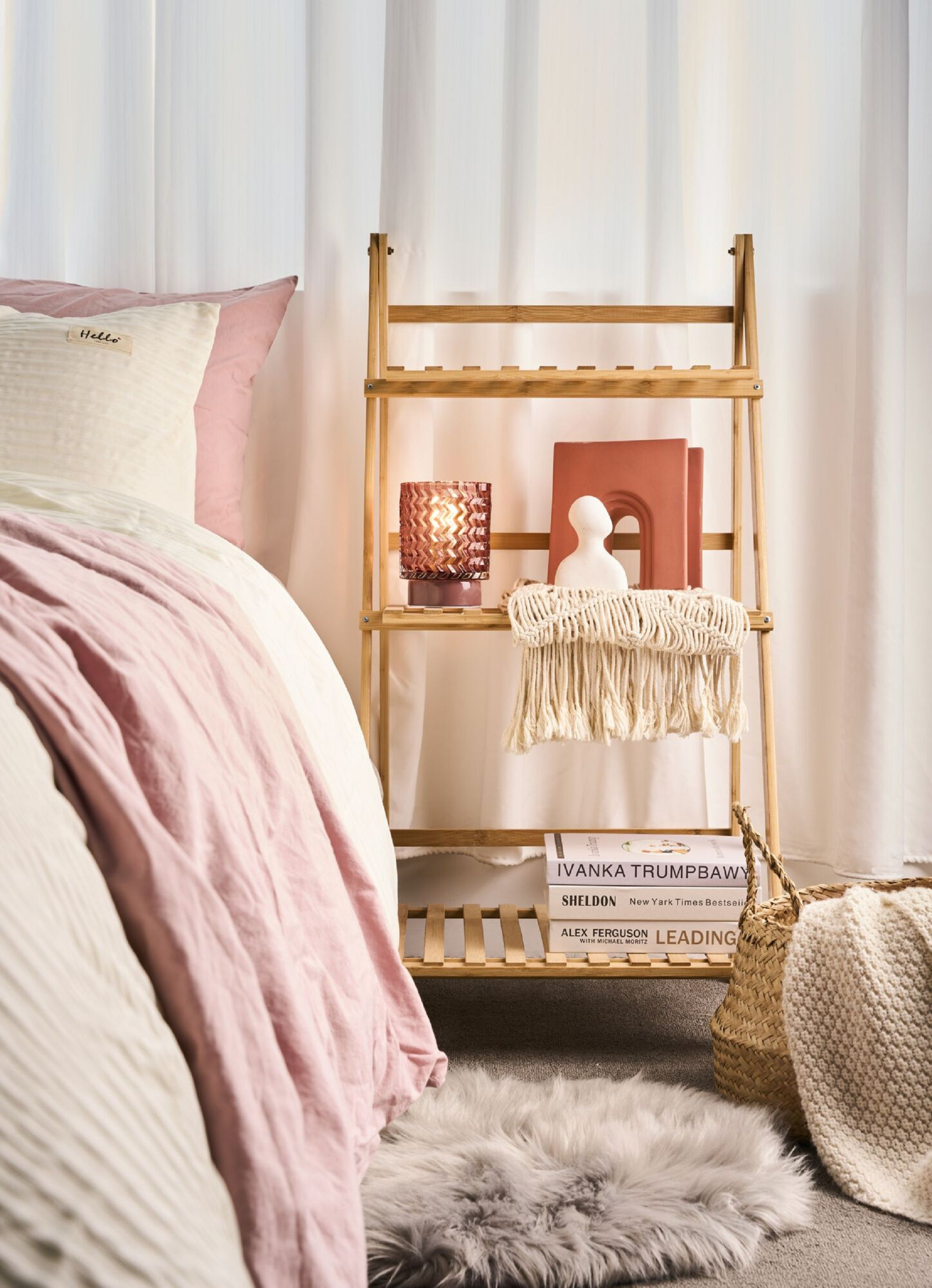 Scena przy łóżku: regał, lampa, książki, poduszki i różowy koc. Białe zasłony.