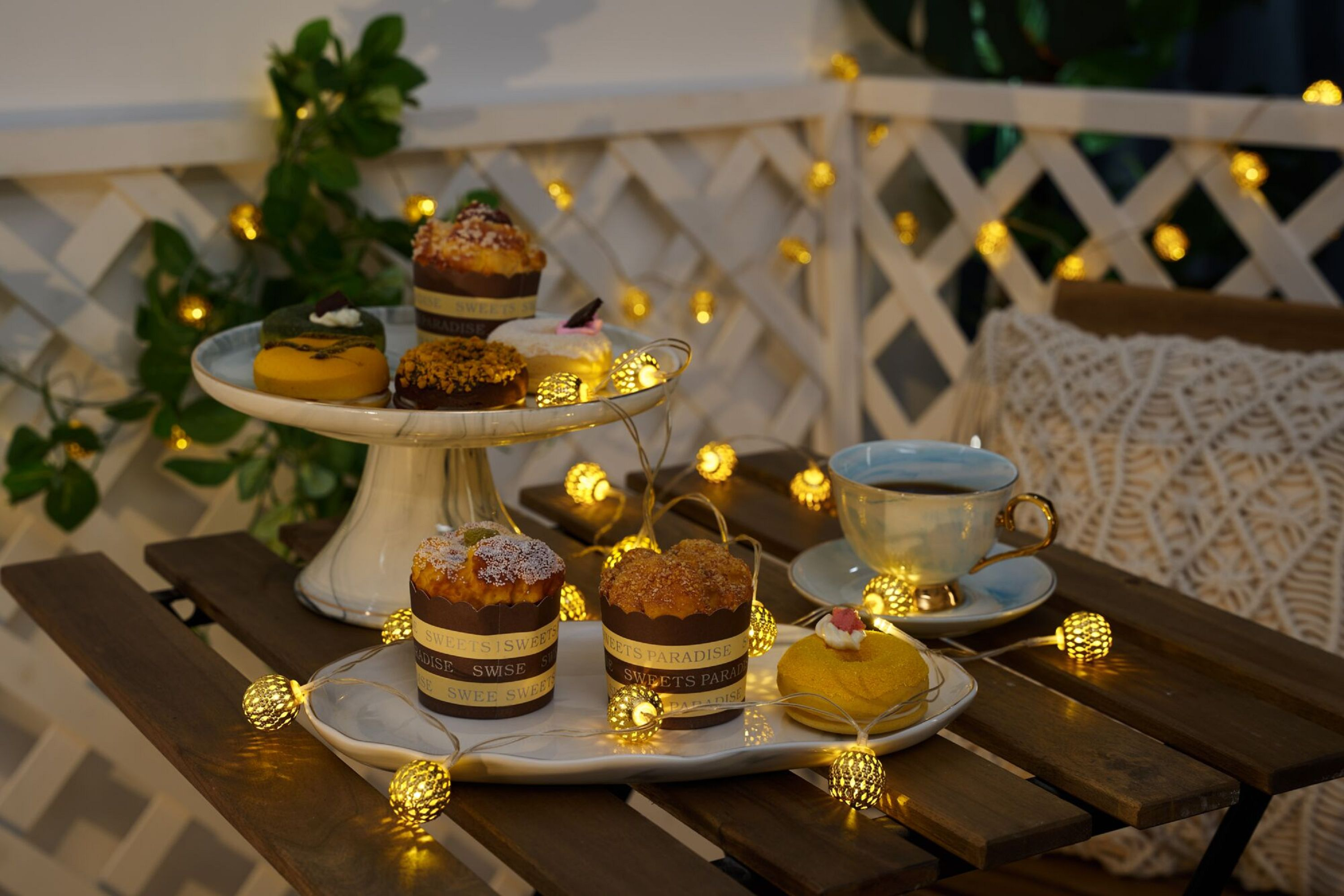 Stół z deserami i światłami. Babeczki na stojaku, filiżanka herbaty i lampki choinkowe oświetlają scenę.
