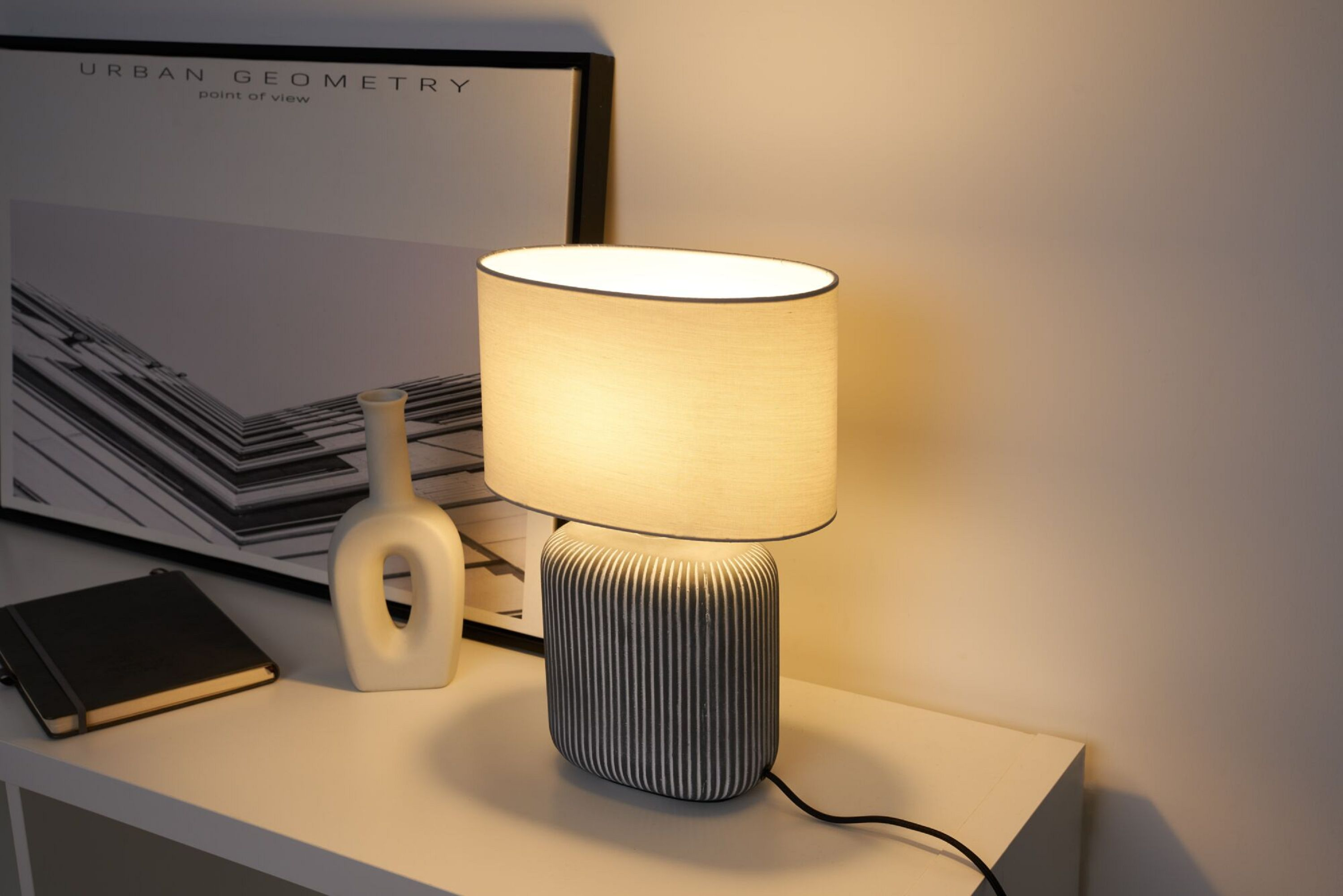 Lampa stołowa z wzorzystą podstawą i owalnym abażurem. Obok obrazu i notatnika.