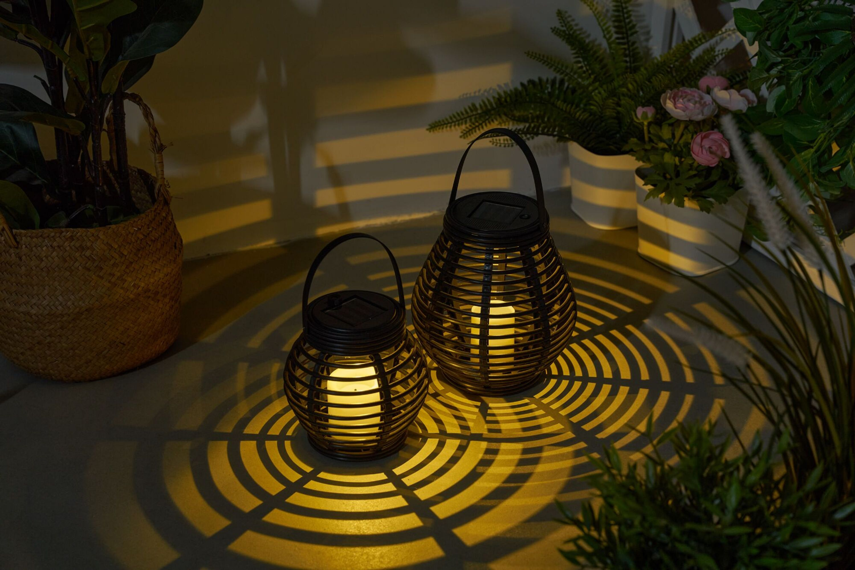 Dwie czarne latarnie z tkanymi bokami, podświetlone, rzucające światło i cień na ziemię z roślinami w tle.