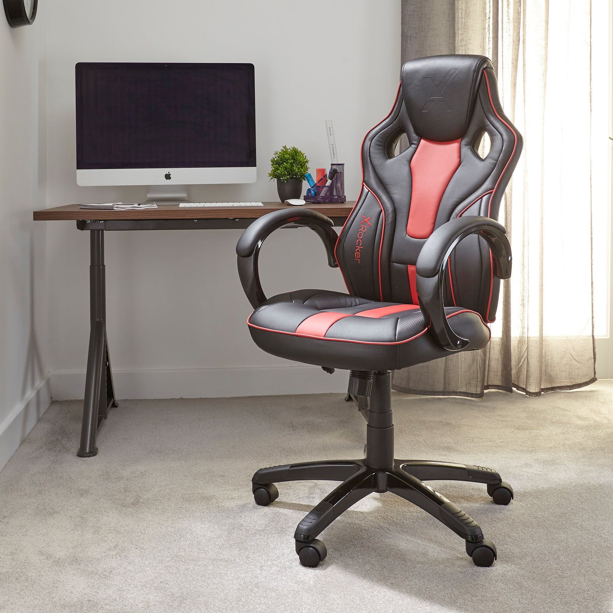 Gaming-Stuhl mit rotem und schwarzem Design, Schreibtisch und Monitor im Hintergrund. Teppichboden.