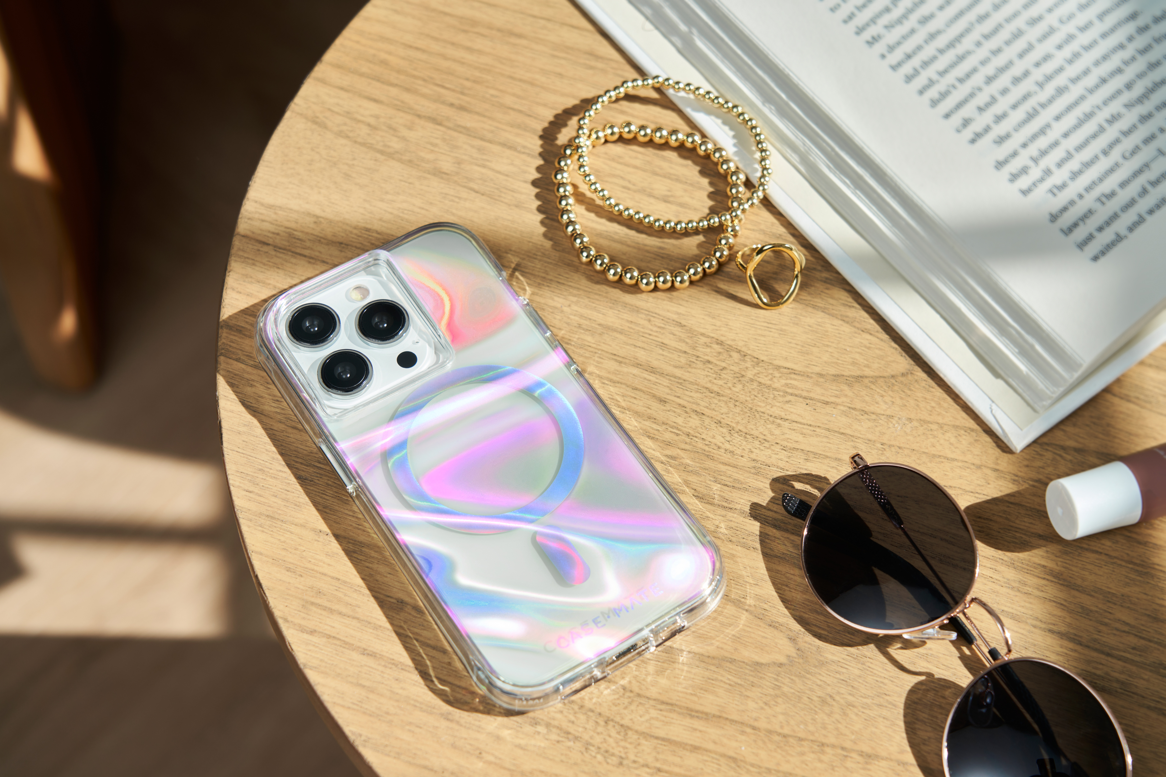 Telefon in holografischer Hülle auf Tisch mit Zubehör: Armband, Sonnenbrille und Lipgloss. Holzoberfläche.