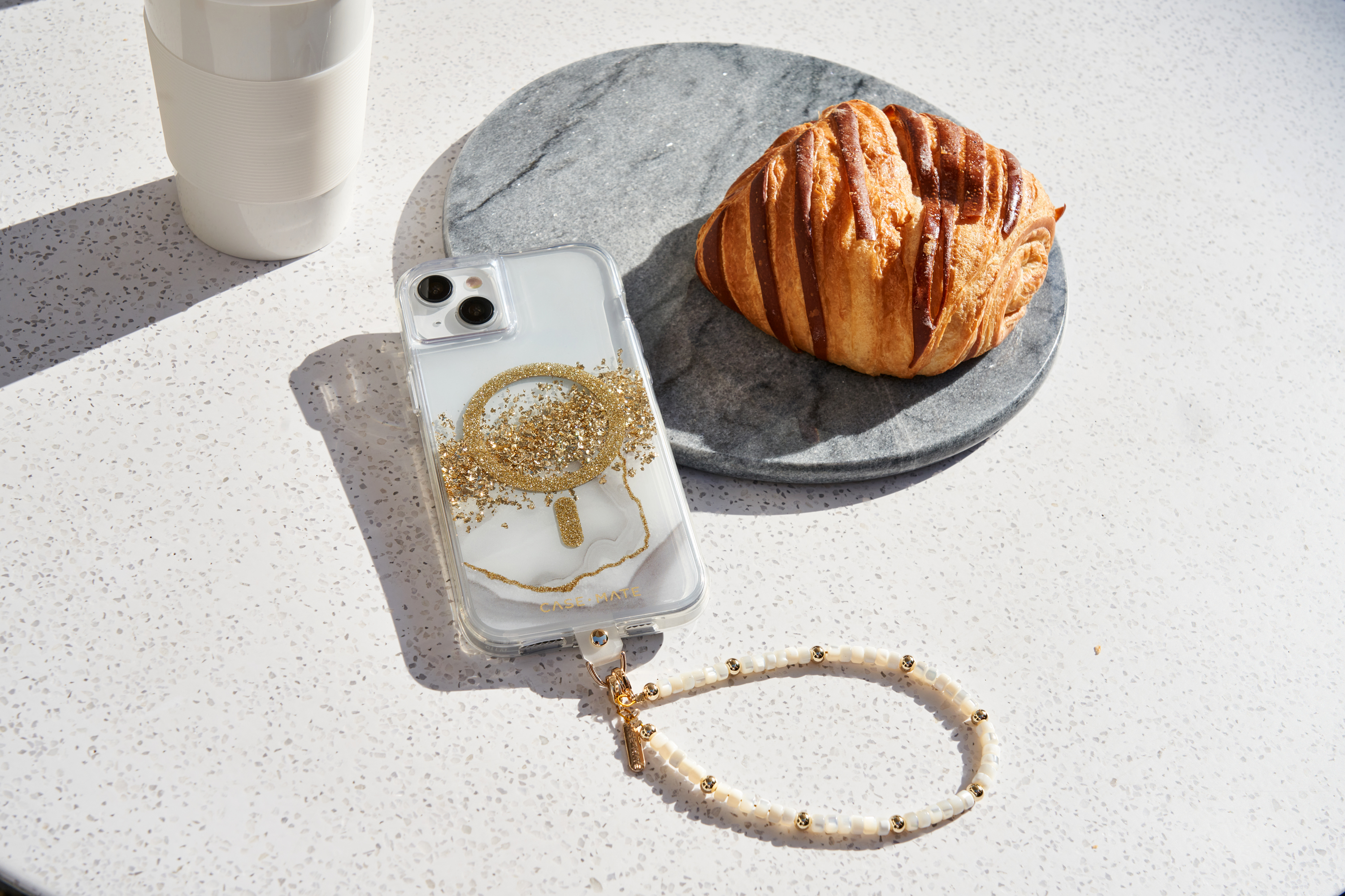 Ein Telefon in einer durchsichtigen Hülle mit goldenen Akzenten. Auf einem Tisch liegen ein Croissant und eine Tasse.