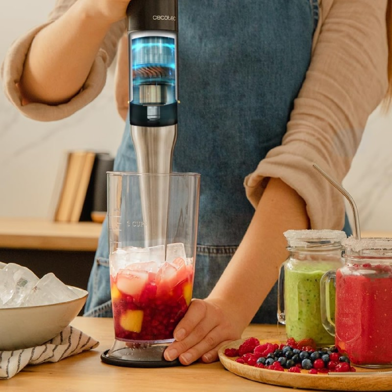 Osoba używająca blendera do przygotowania koktajlu. Szklany słoik, lód i owoce na pierwszym planie. Drewniany stół.