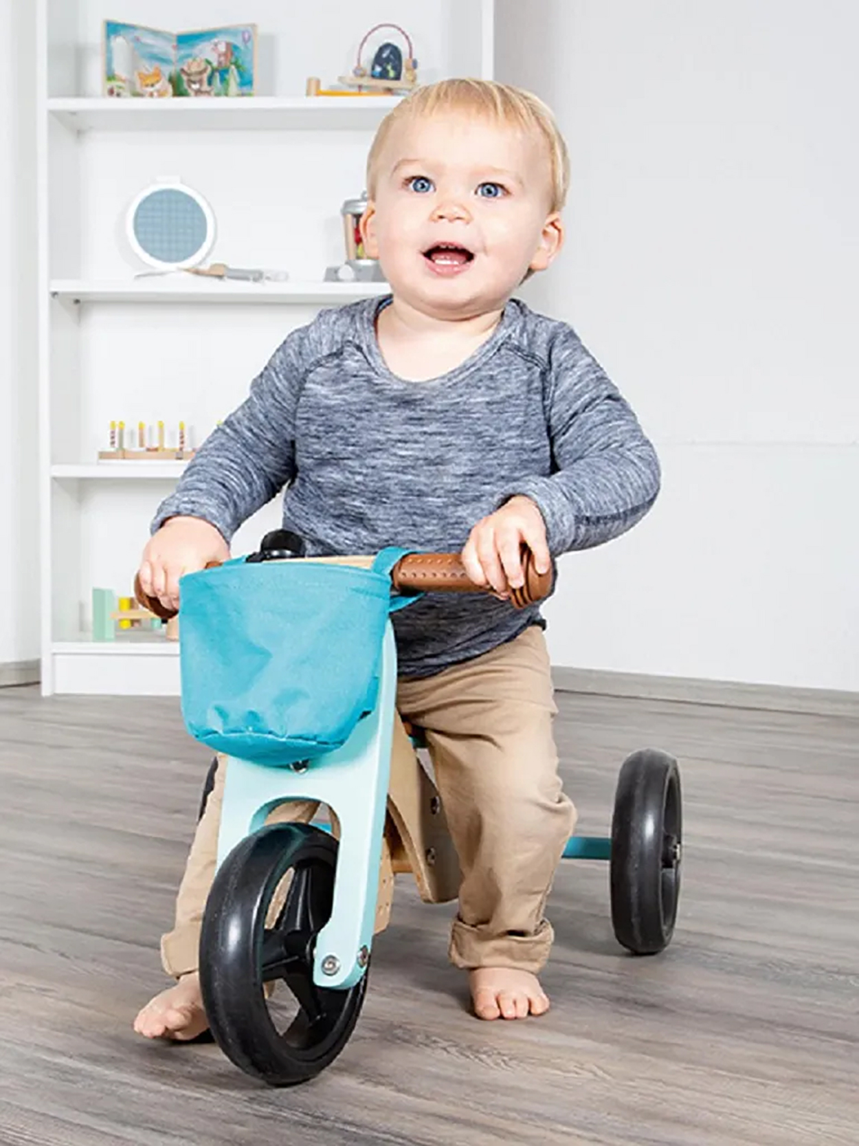 Un bambino su un triciclo blu sorride. Ha un cestino e pantaloni marroni. Una mensola è dietro.