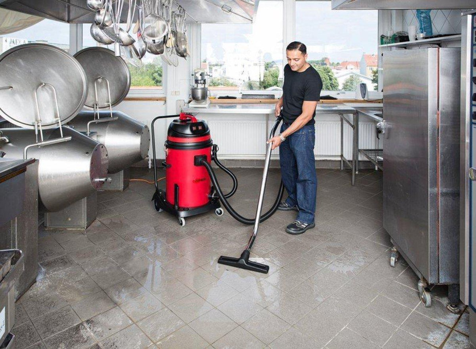 Un uomo aspira il pavimento di una cucina. Si vedono un aspirapolvere rosso e attrezzature in acciaio inox.