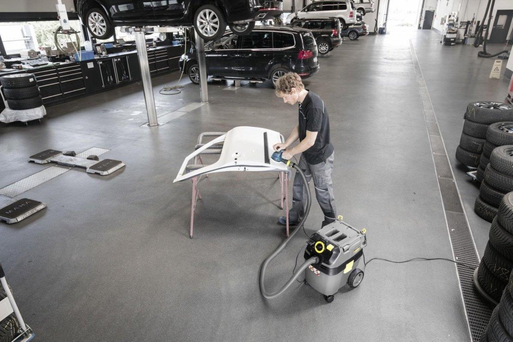 Osoba szlifująca maskę samochodu w garażu z odkurzaczem w pobliżu.