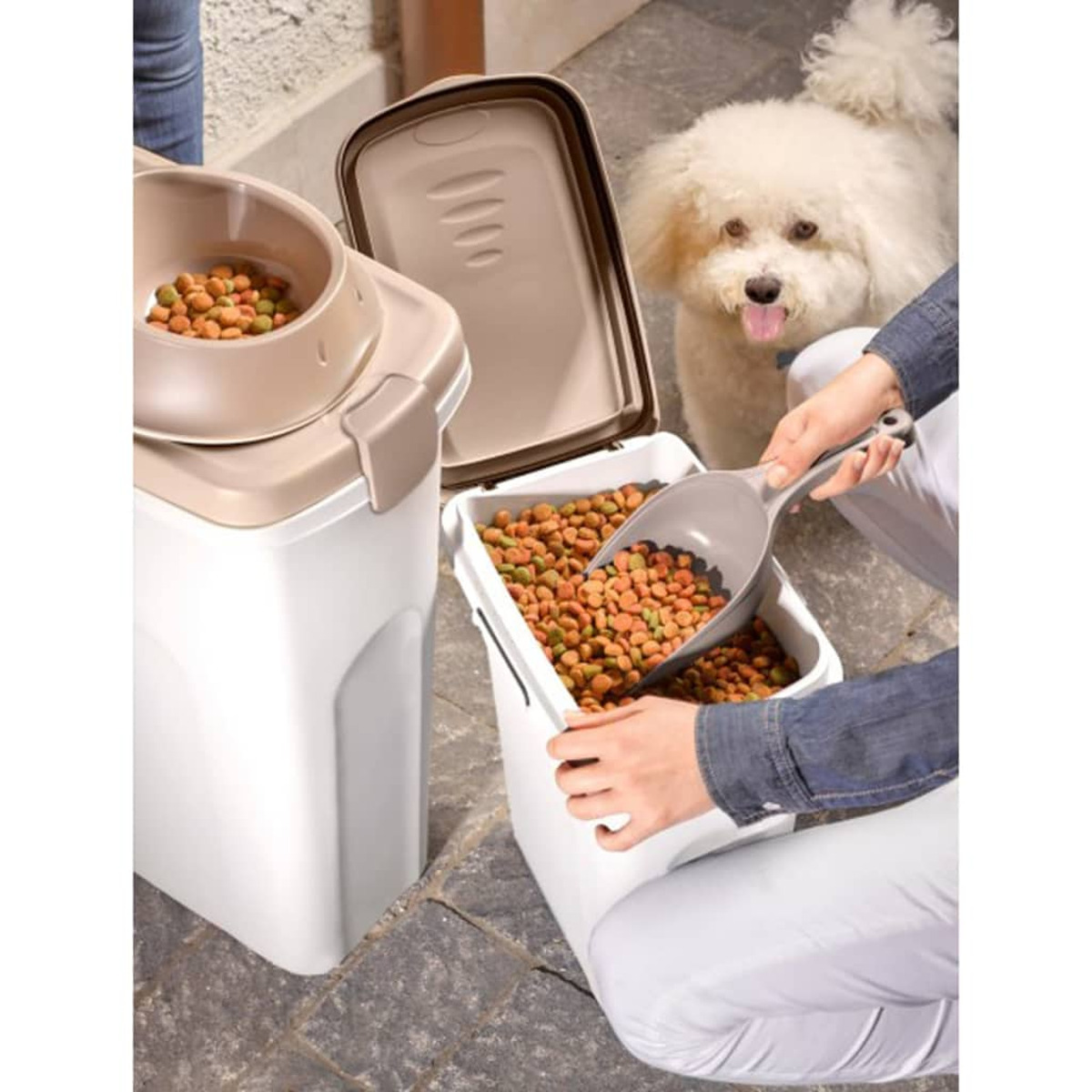 Eine Person füllt einen Behälter mit Hundefutter, während ein Hund aufgeregt zuschaut. Das Hundefutter ist sichtbar.