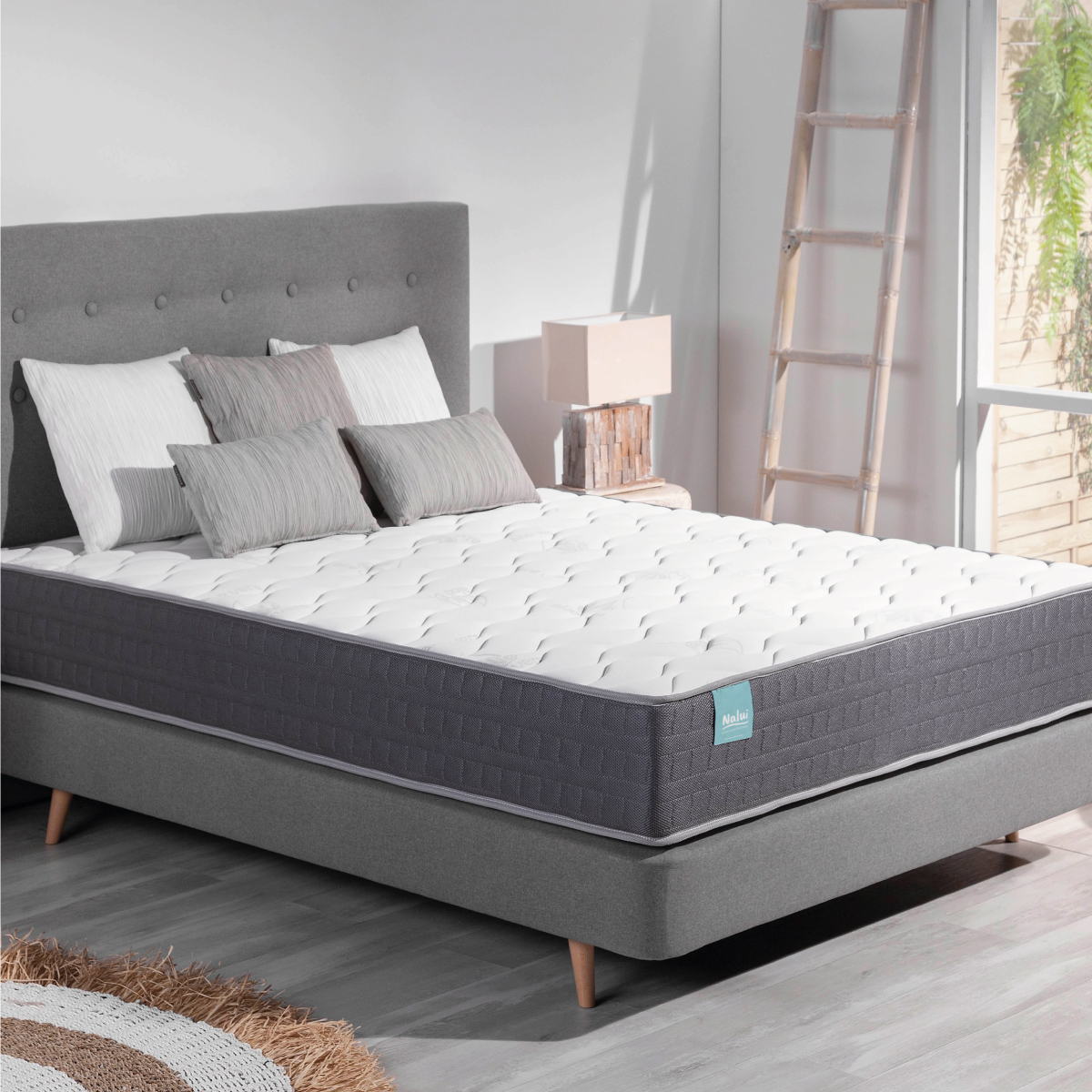 Una cama con cabecero gris, almohadas blancas y un colchón blanco y gris. Hay una escalera de madera.