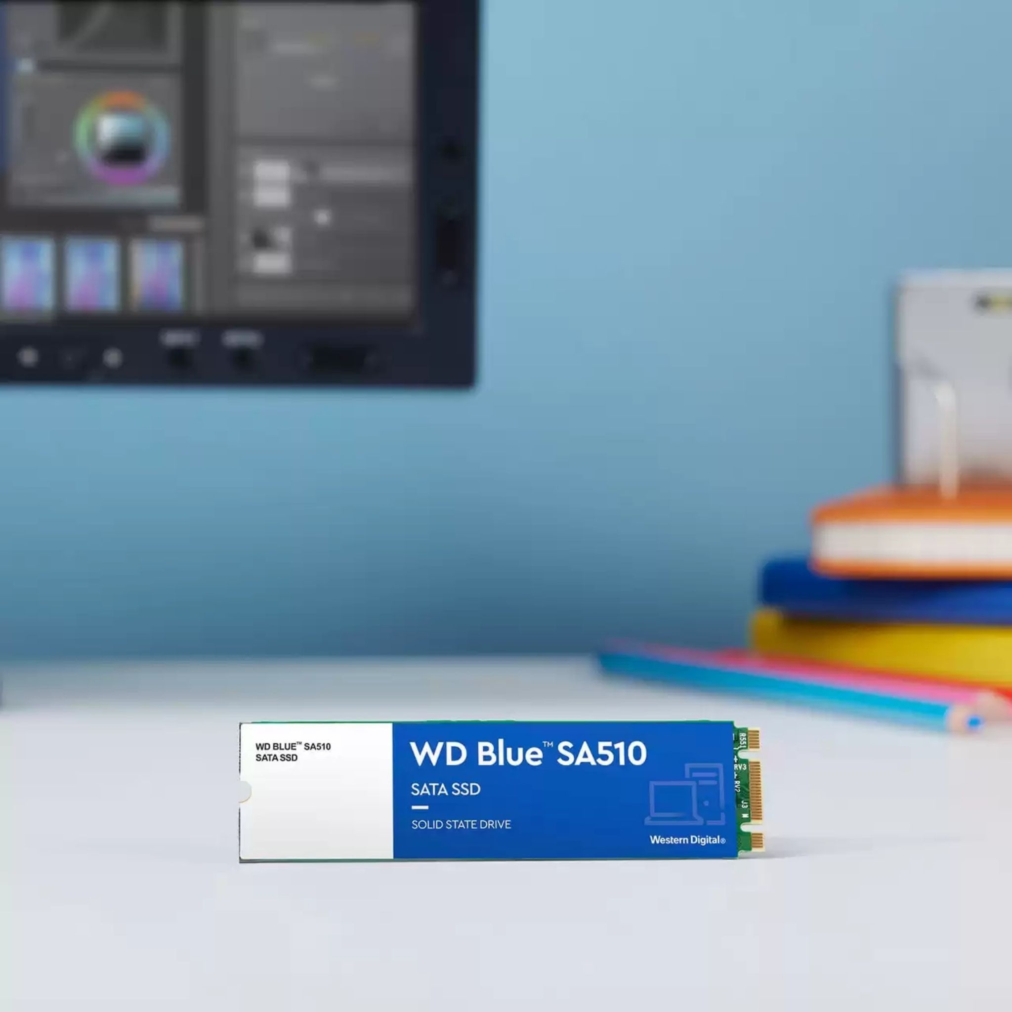 Dysk SSD z niebieską etykietą, WD Blue, na białym biurku z kolorowymi książkami i monitorem.