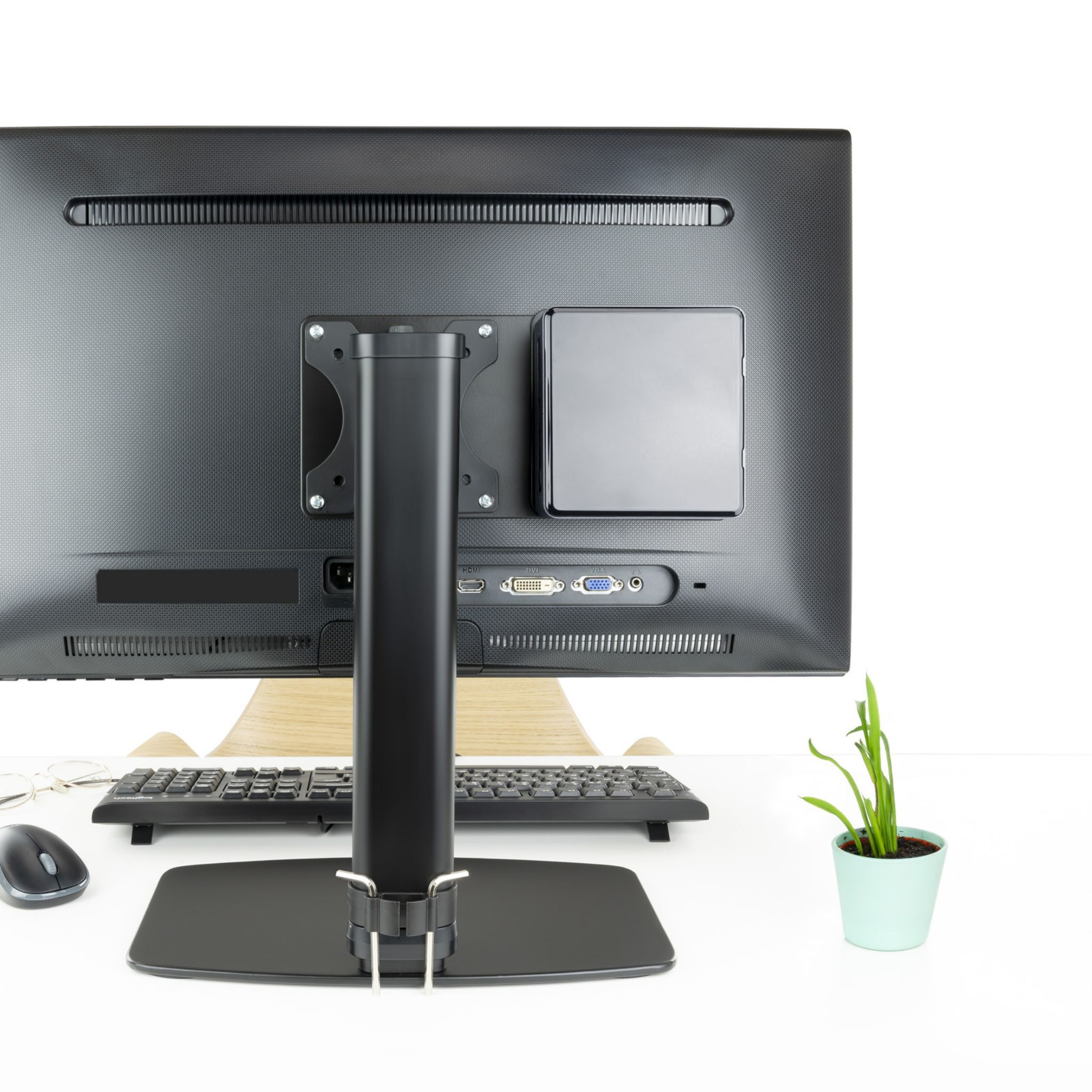 Parte trasera del monitor con ordenador. Teclado, ratón y planta en un escritorio blanco.