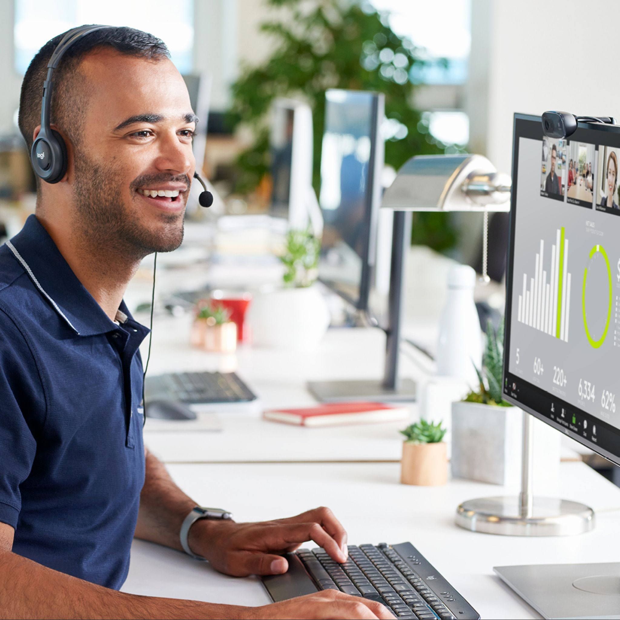 Mann mit Headset, lächelnd, arbeitet am Computer mit Monitor, der Diagramme und Videoanrufe zeigt.
