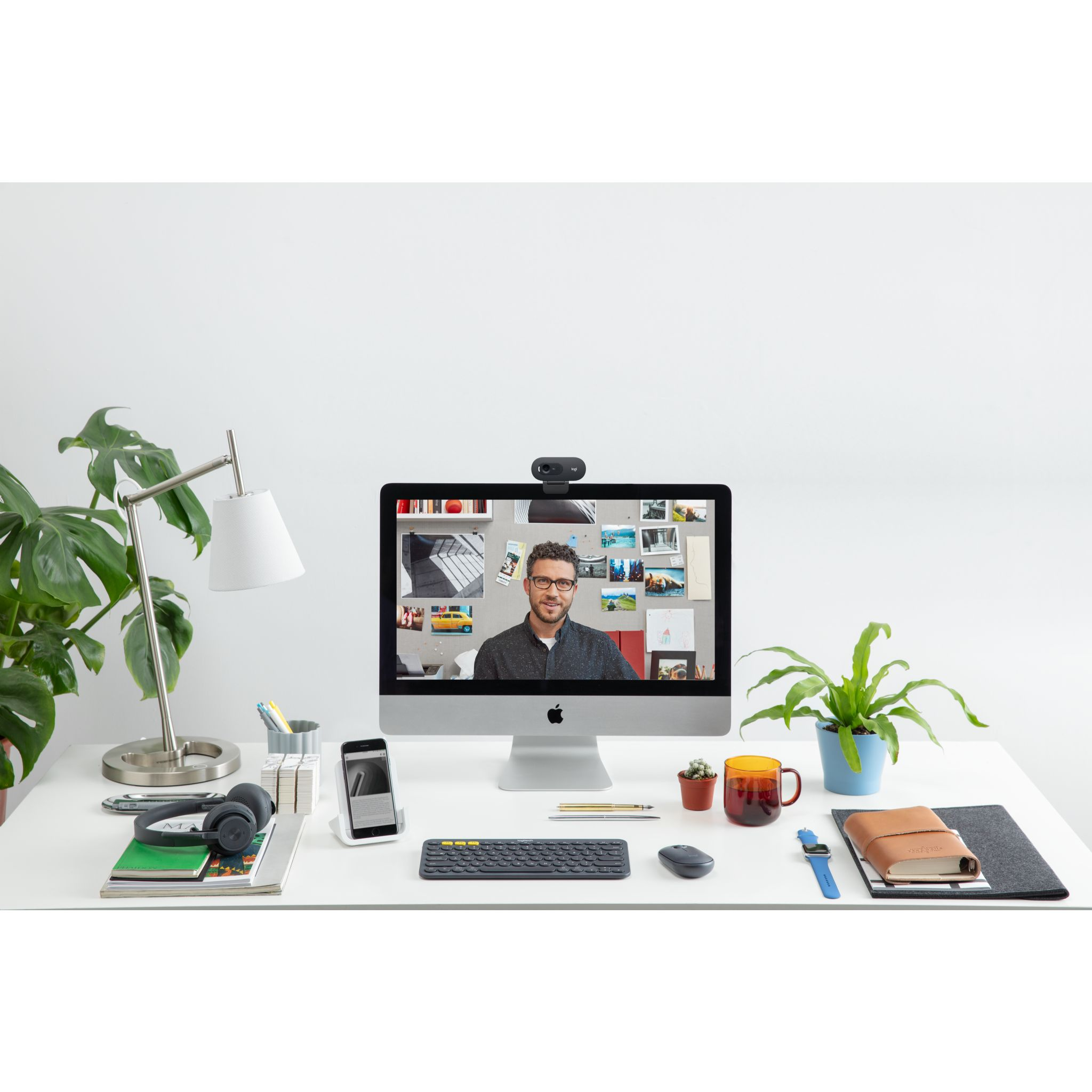 Konfiguracja biurka z osobą na ekranie, komputerem, telefonem, akcesoriami i rośliną.