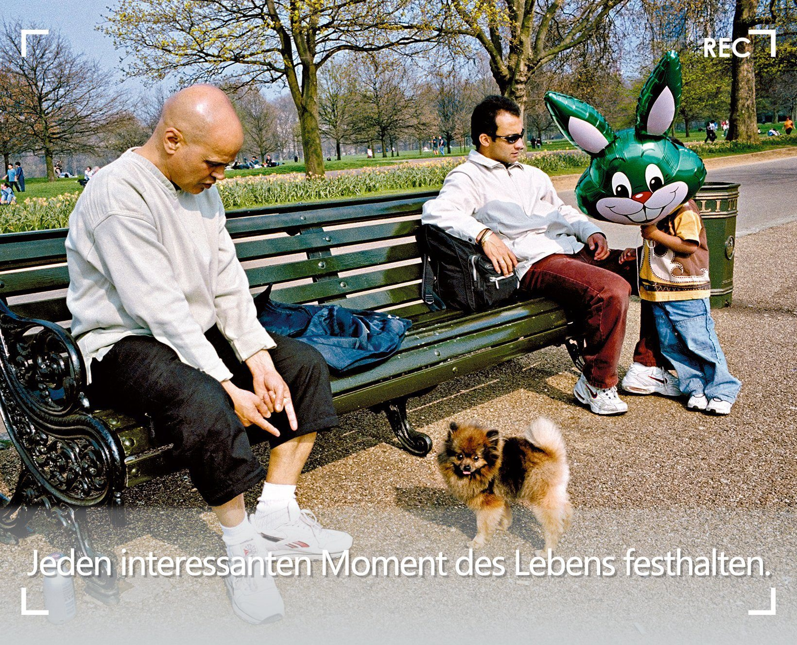 Zwei Männer und ein Kind mit einem Hasenballon auf einer Parkbank, mit einem Hund.