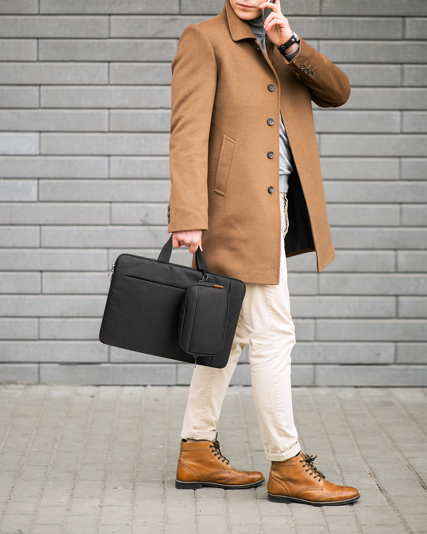 Mann mit Mantel hält schwarze Tasche, telefoniert. Steht auf einem Ziegelweg.