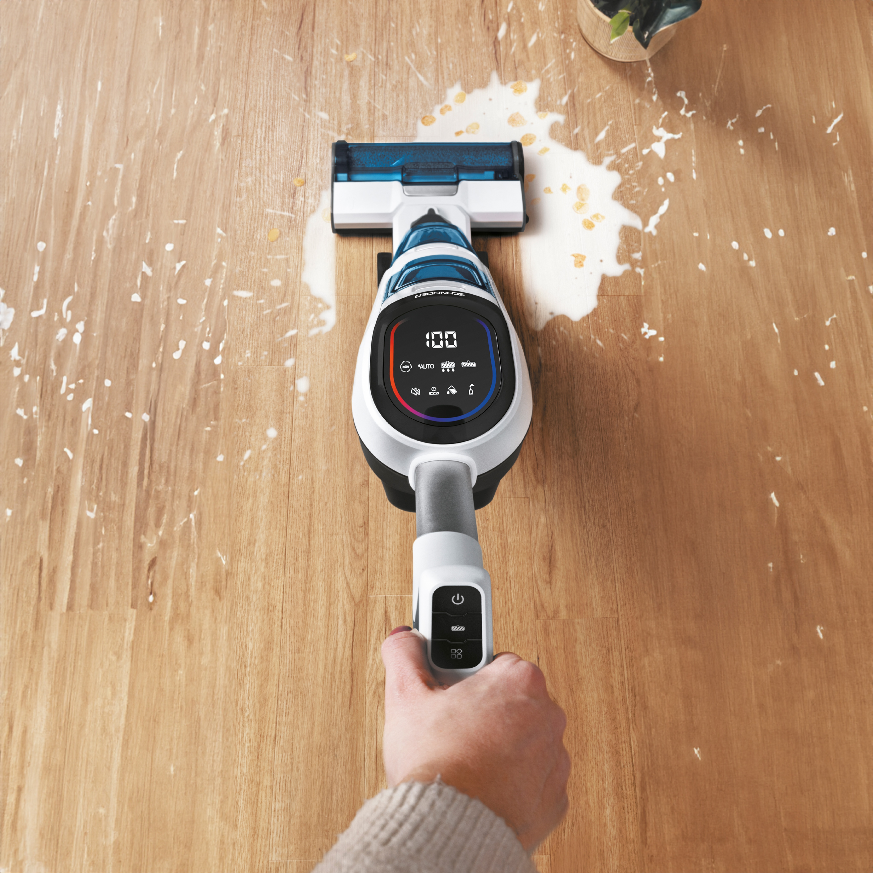 Un aspirapolvere portatile pulisce un pavimento in legno con liquido versato e macchie bianche.