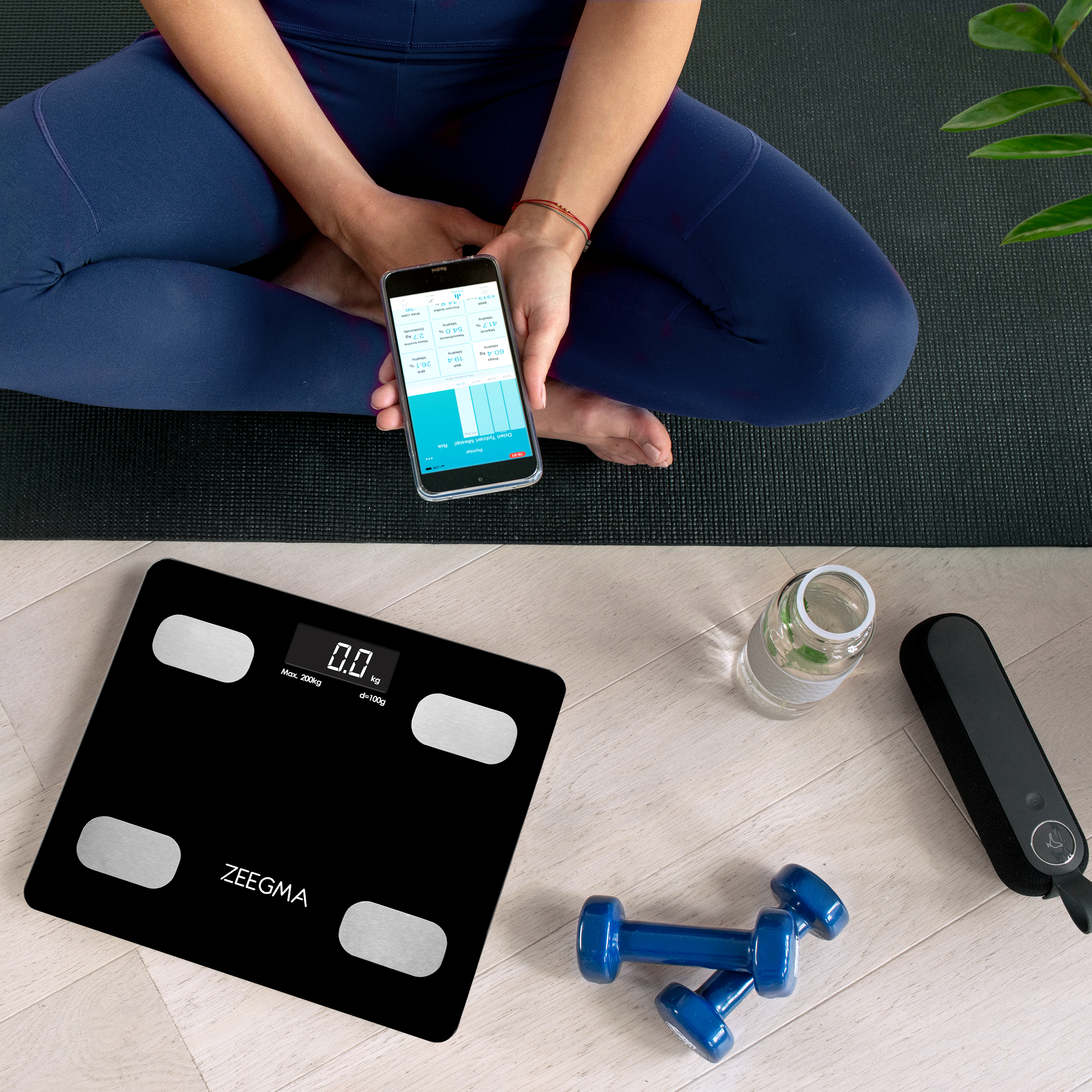 Kobieta w niebieskich legginsach siedzi, trzymając telefon. Z przodu waga, hantle i butelka wody.