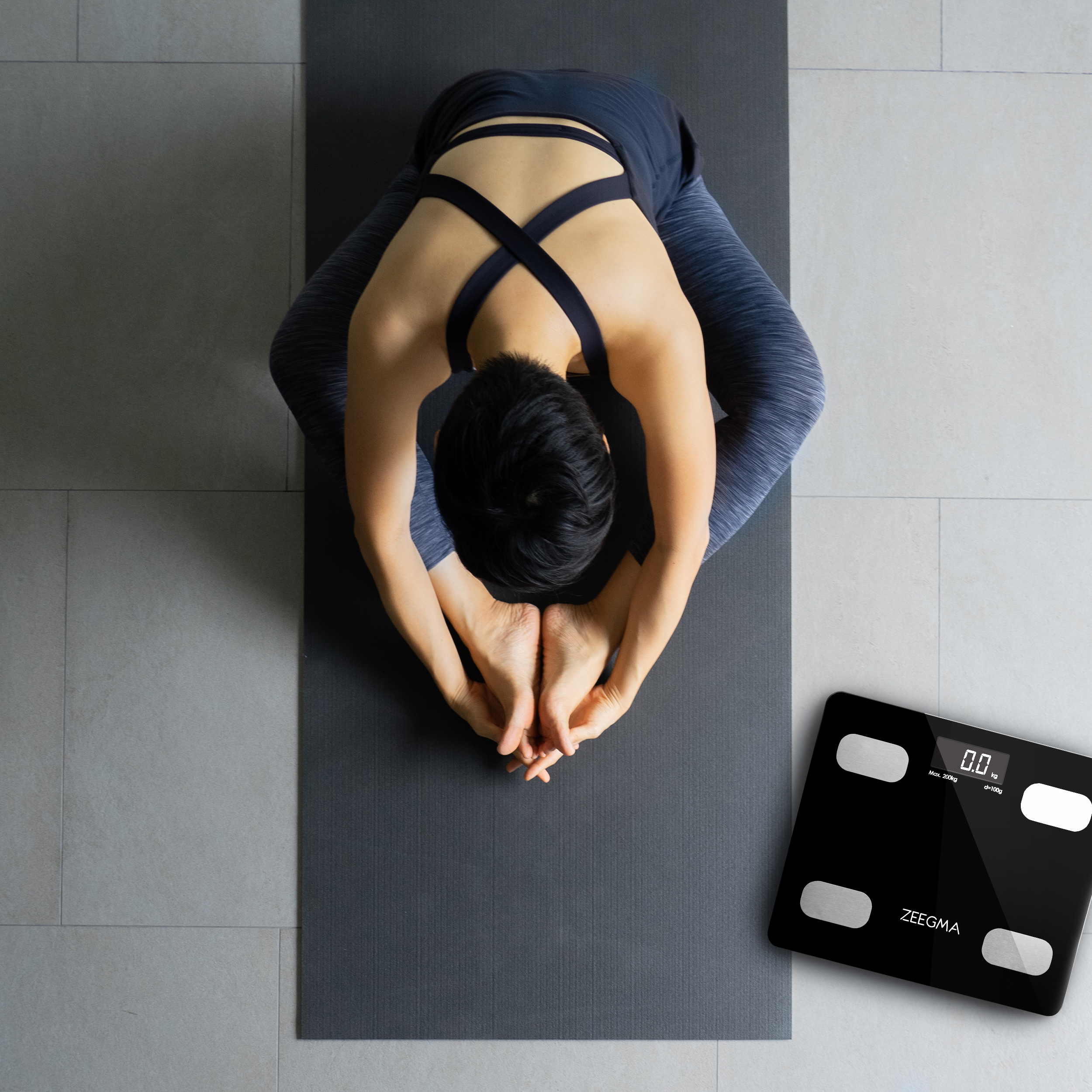 Kobieta rozciąga się na macie do jogi, obok czarna waga. Podłoga jest szara.