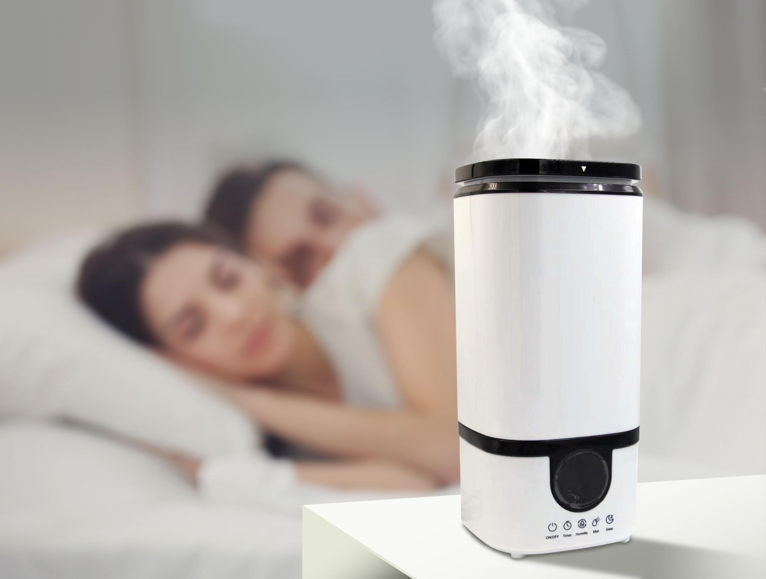 Un humidificador blanco, con vapor ascendente, sobre una mesa en un dormitorio, con una pareja borrosa.