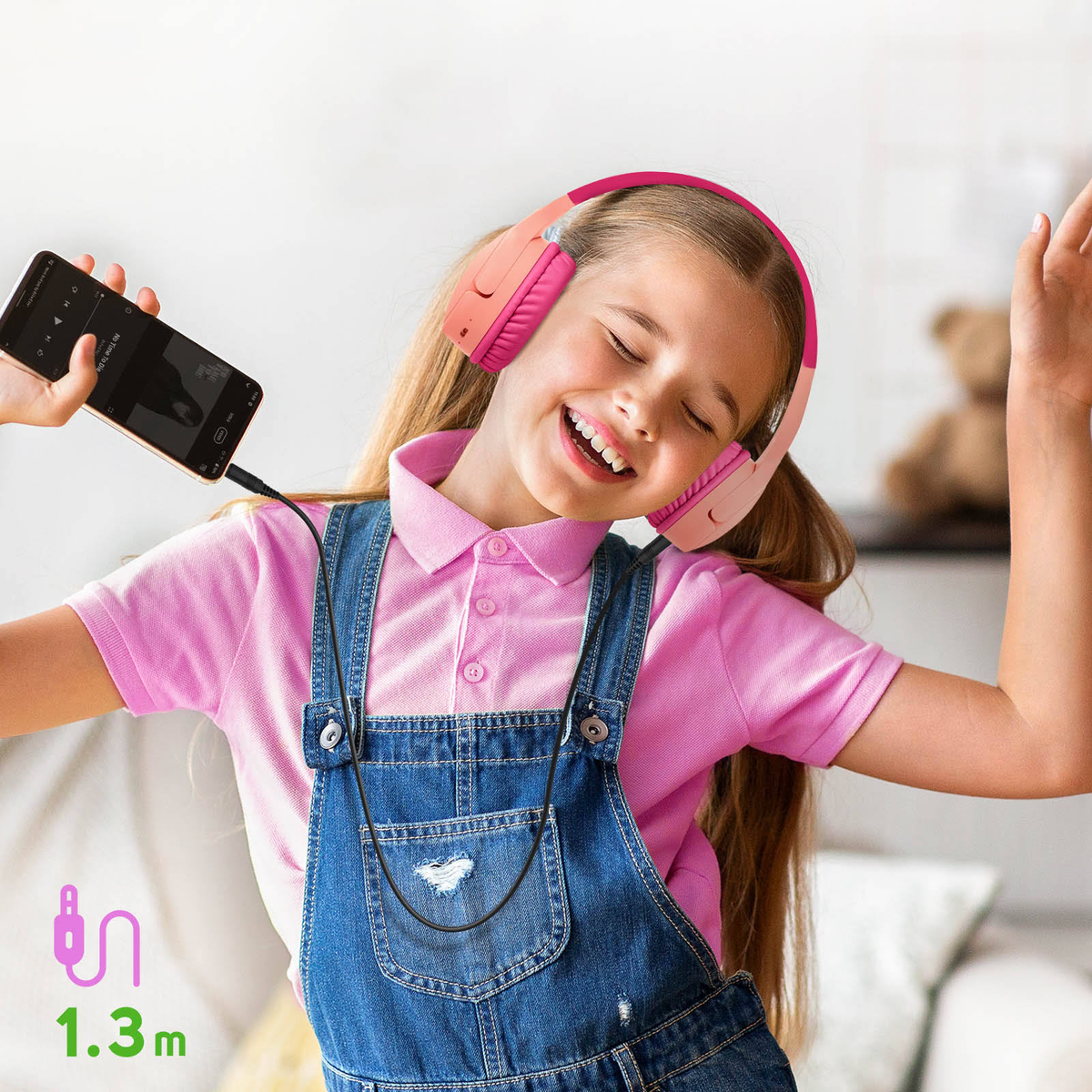 Ragazza con cuffie rosa e tuta di jeans, ascolta musica su un telefono. Sorridente.