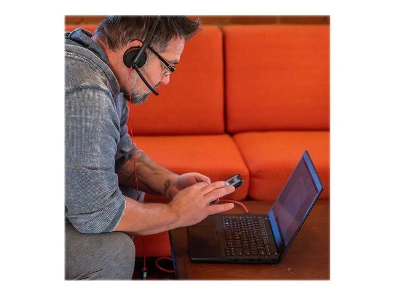 Mężczyzna używający laptopa i smartfona, z zestawem słuchawkowym, siedzący na kanapie.