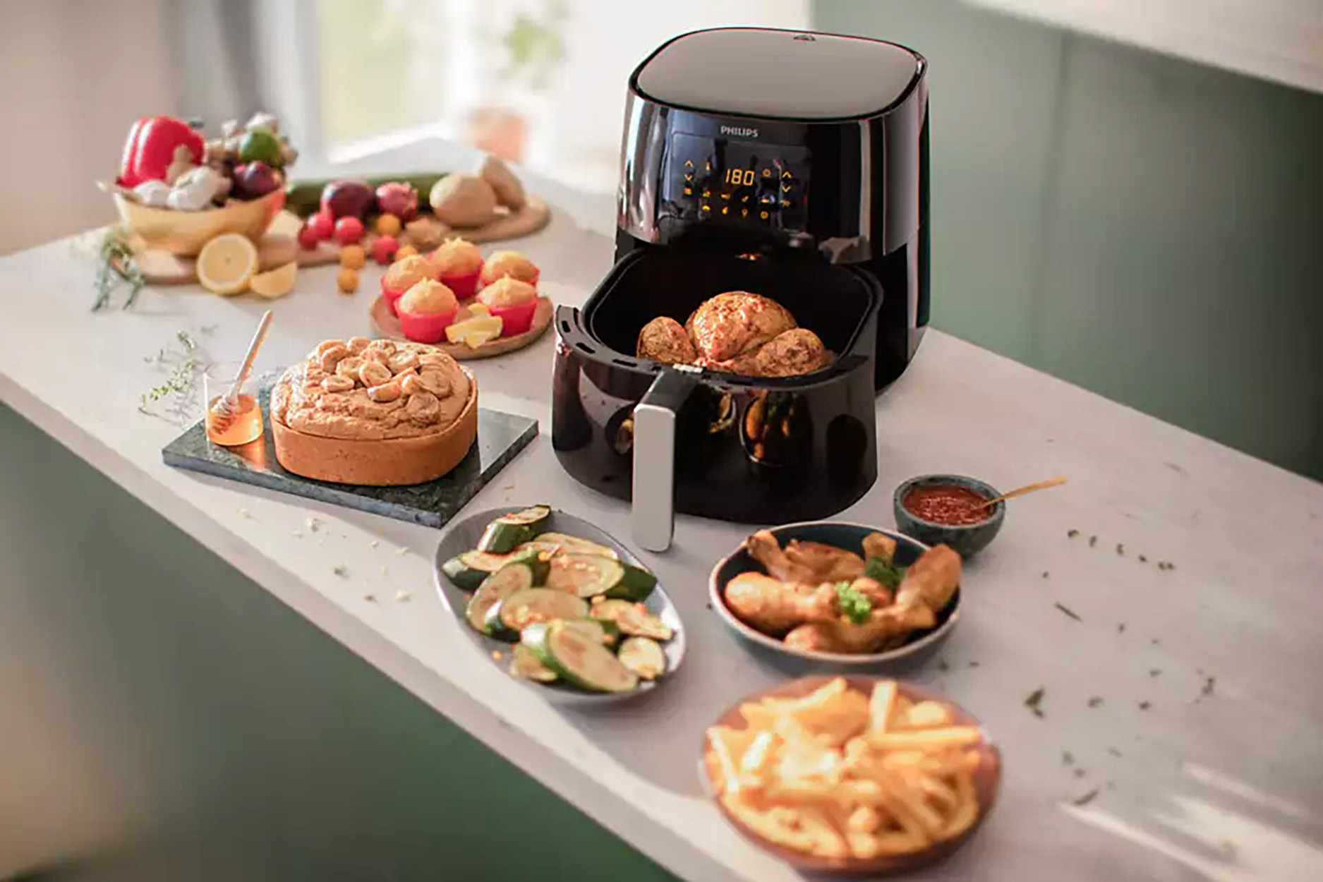 Zwarte airfryer met gekookt voedsel en ingrediënten eromheen. Bevat een cake, groenten en friet.