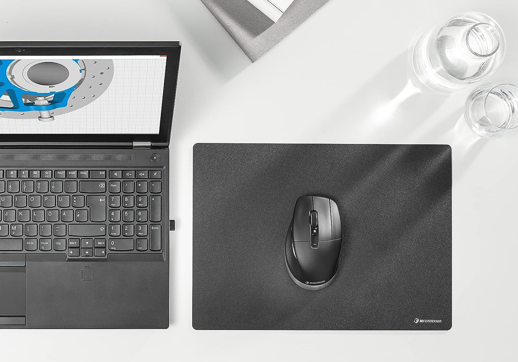 Laptop zeigt CAD-Design. Maus und Mauspad auf weißem Schreibtisch. Ein Wasserglas ist sichtbar.