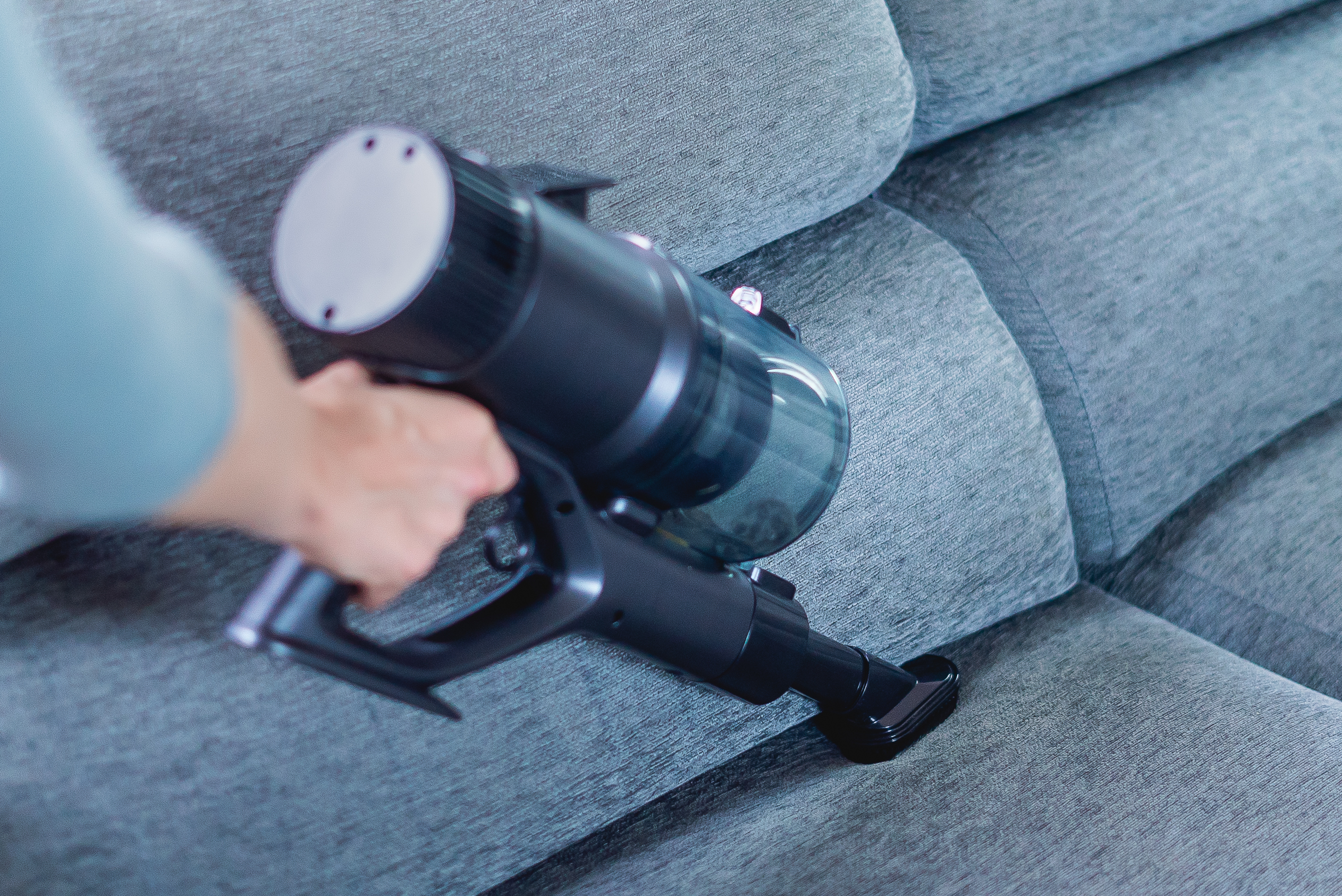 Eine Person saugt mit einem Handstaubsauger ein graues Sofa ab.