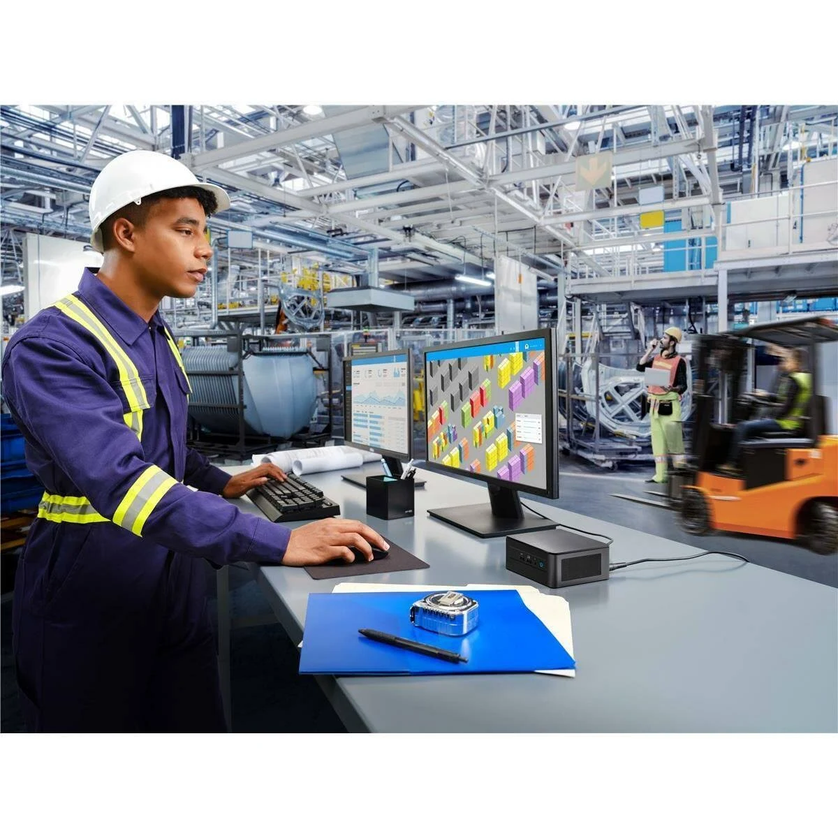 Un lavoratore a una scrivania con doppi monitor, tastiera e accessori in un ambiente industriale.