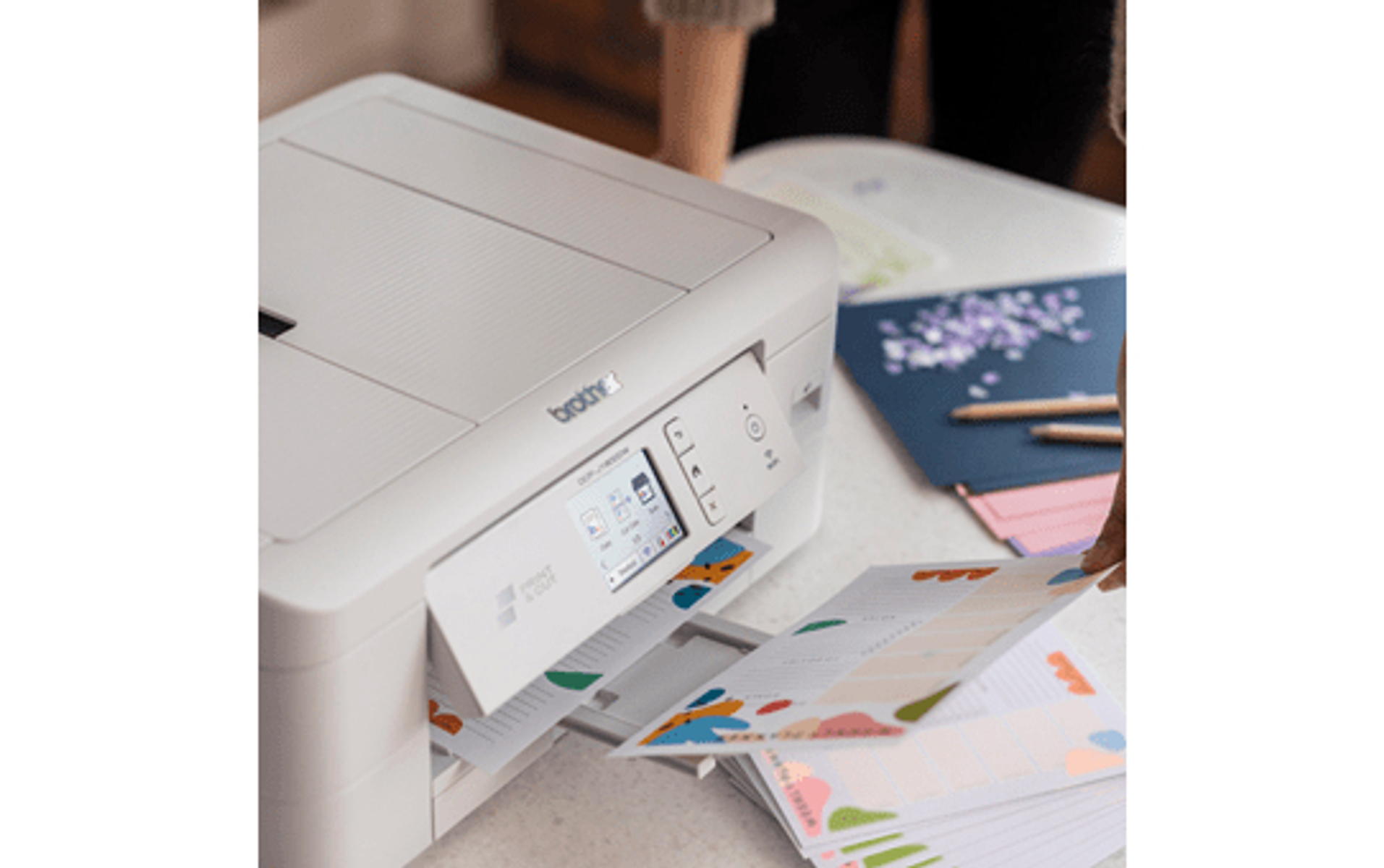 Biała drukarka drukuje kolorowe papiery. Widoczna dłoń osoby. Na stole inne papiery i ołówki.