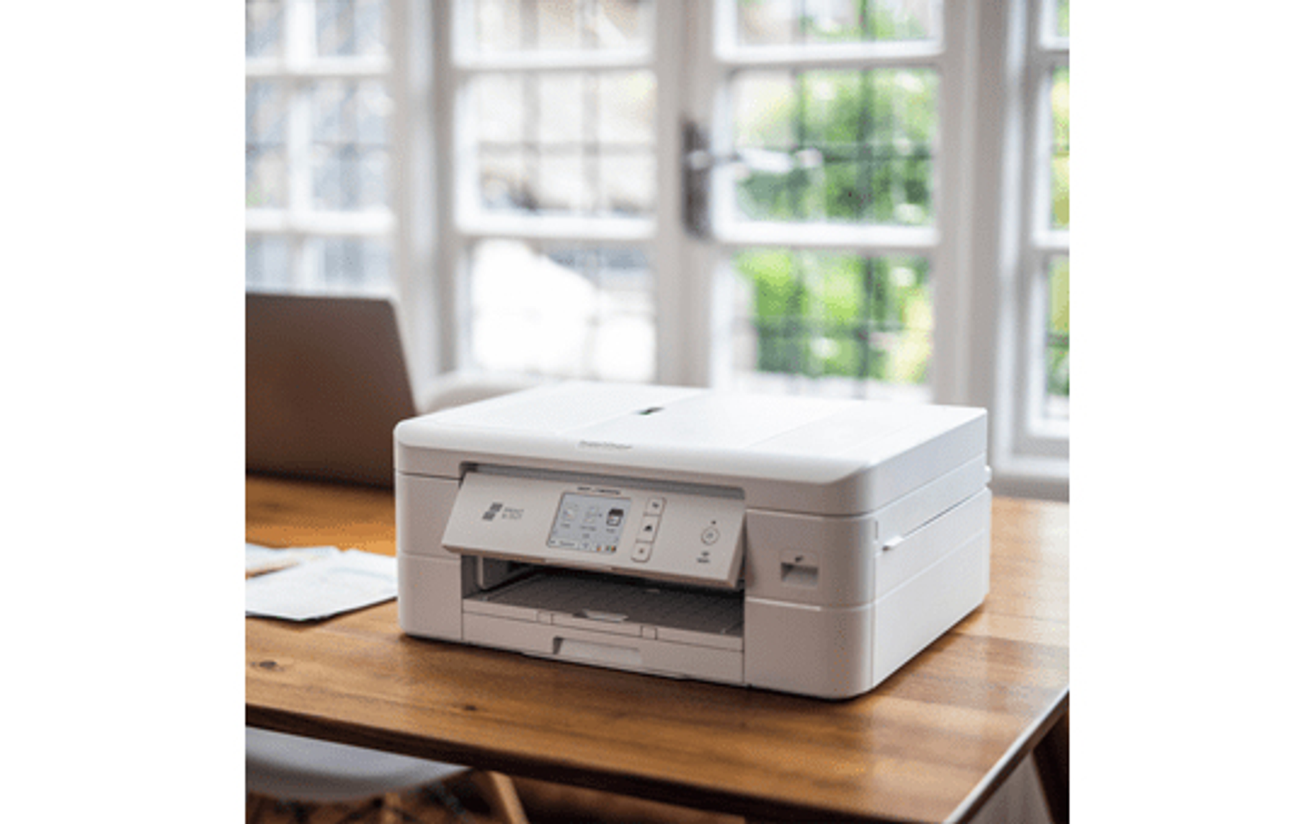 Biała drukarka na drewnianym biurku. Tło przedstawia okna i rozmyty widok natury.