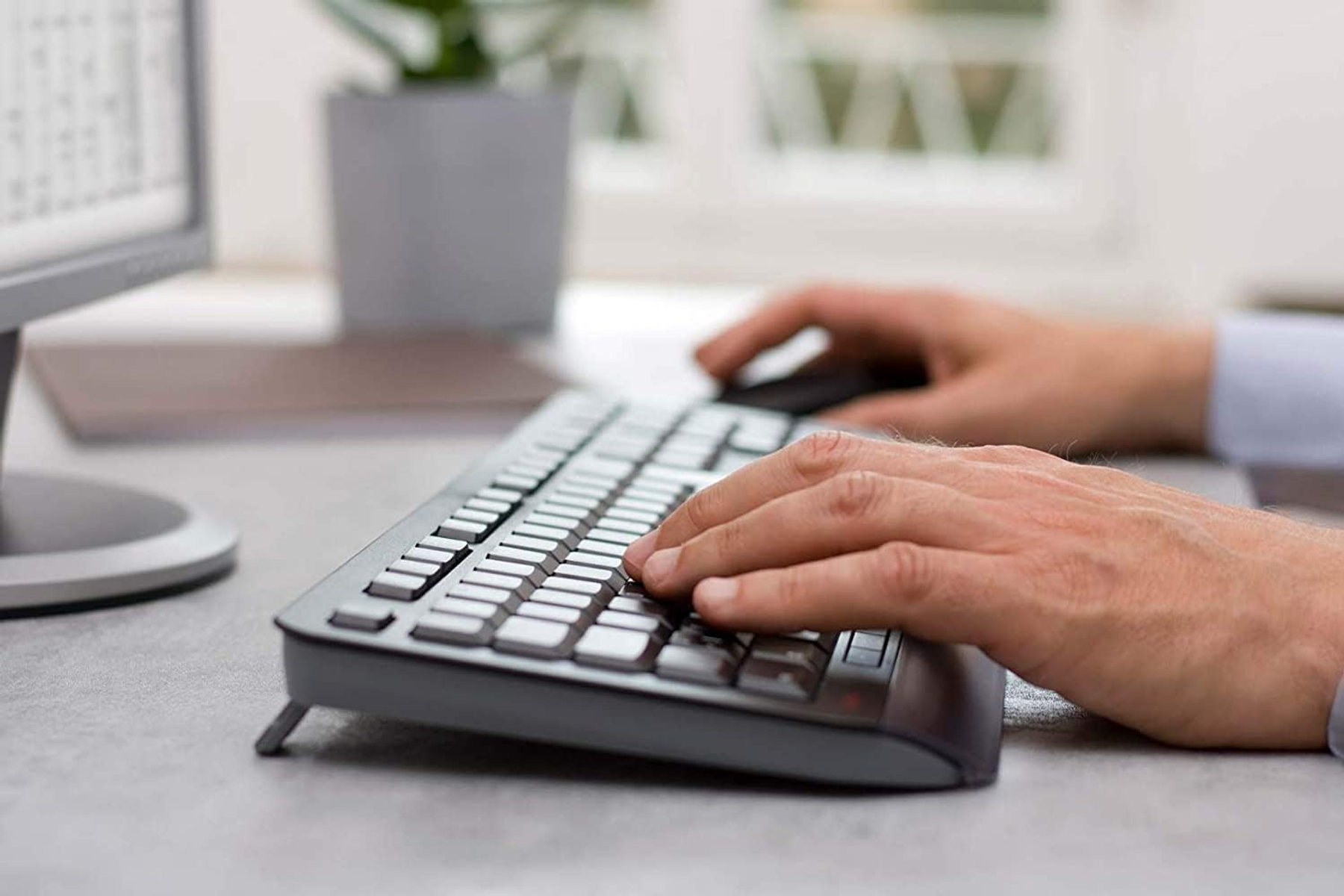 Ręce piszące na klawiaturze, z myszą w pobliżu. W tle monitor i biurko.