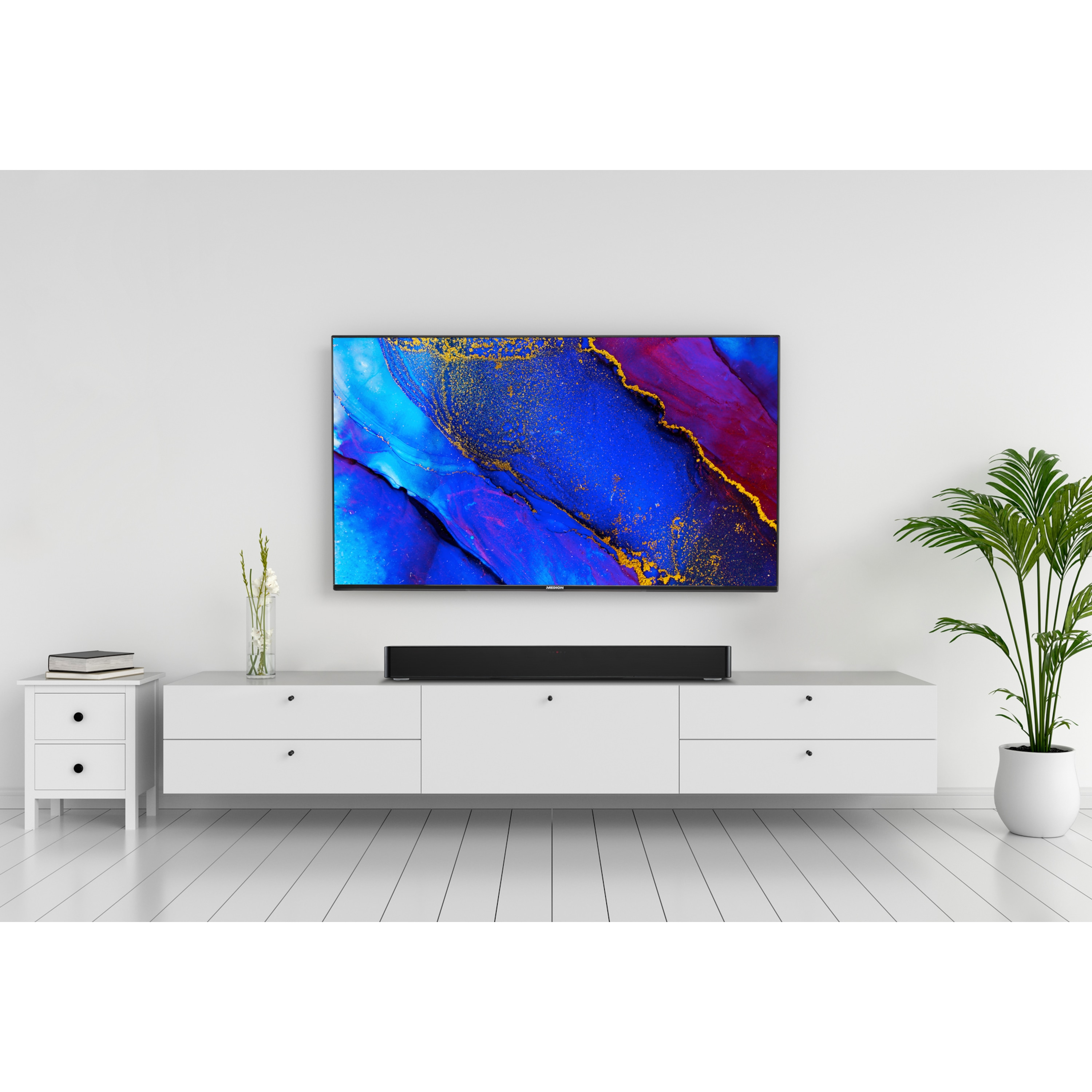 Nowoczesny salon z telewizorem nad białą szafką i soundbarem. Obecna jest roślina i dekoracje.