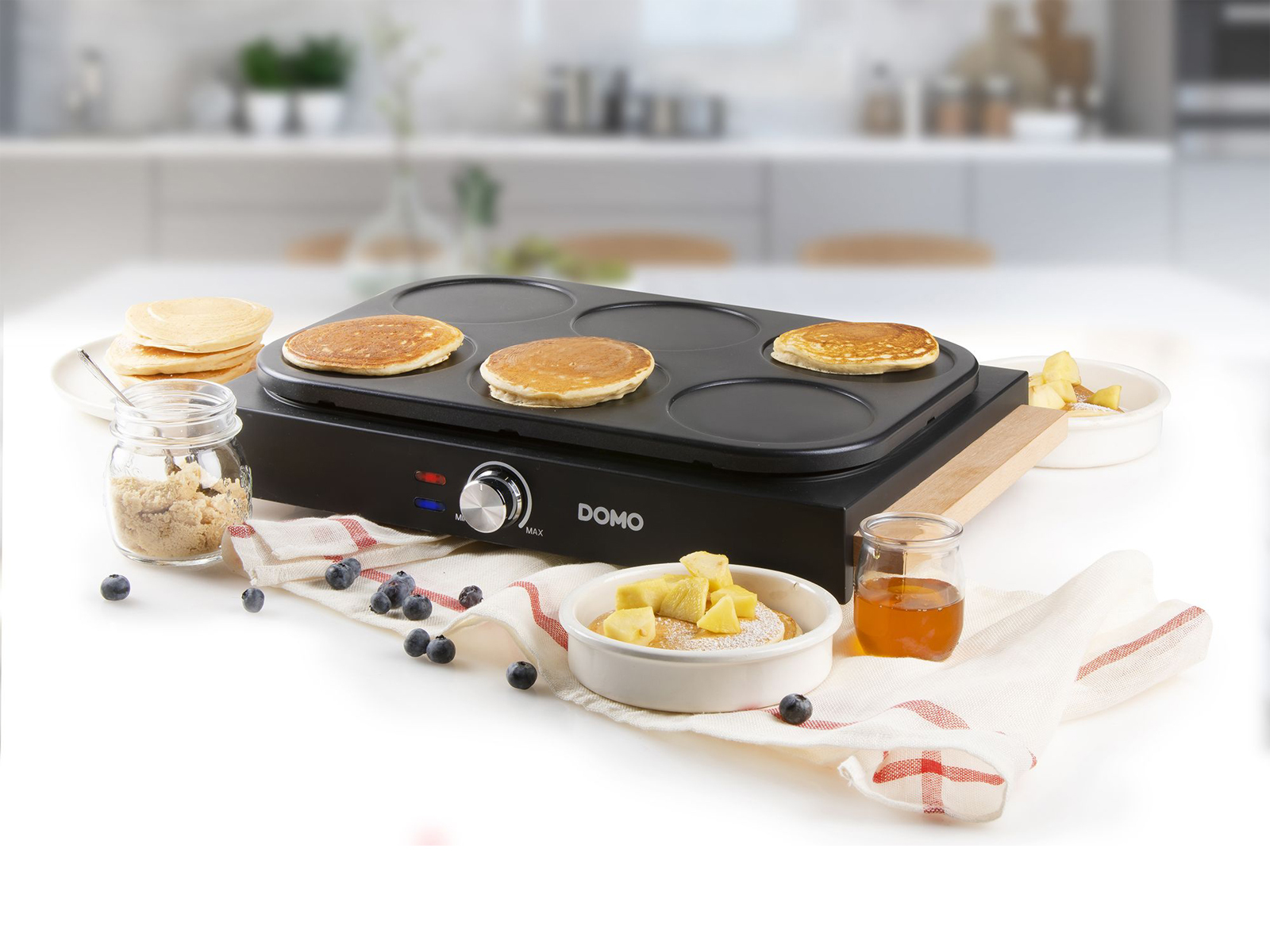 Ein schwarzer Pfannkuchenmacher mit Pfannkuchen, Obst und Toppings auf einer weißen Oberfläche.