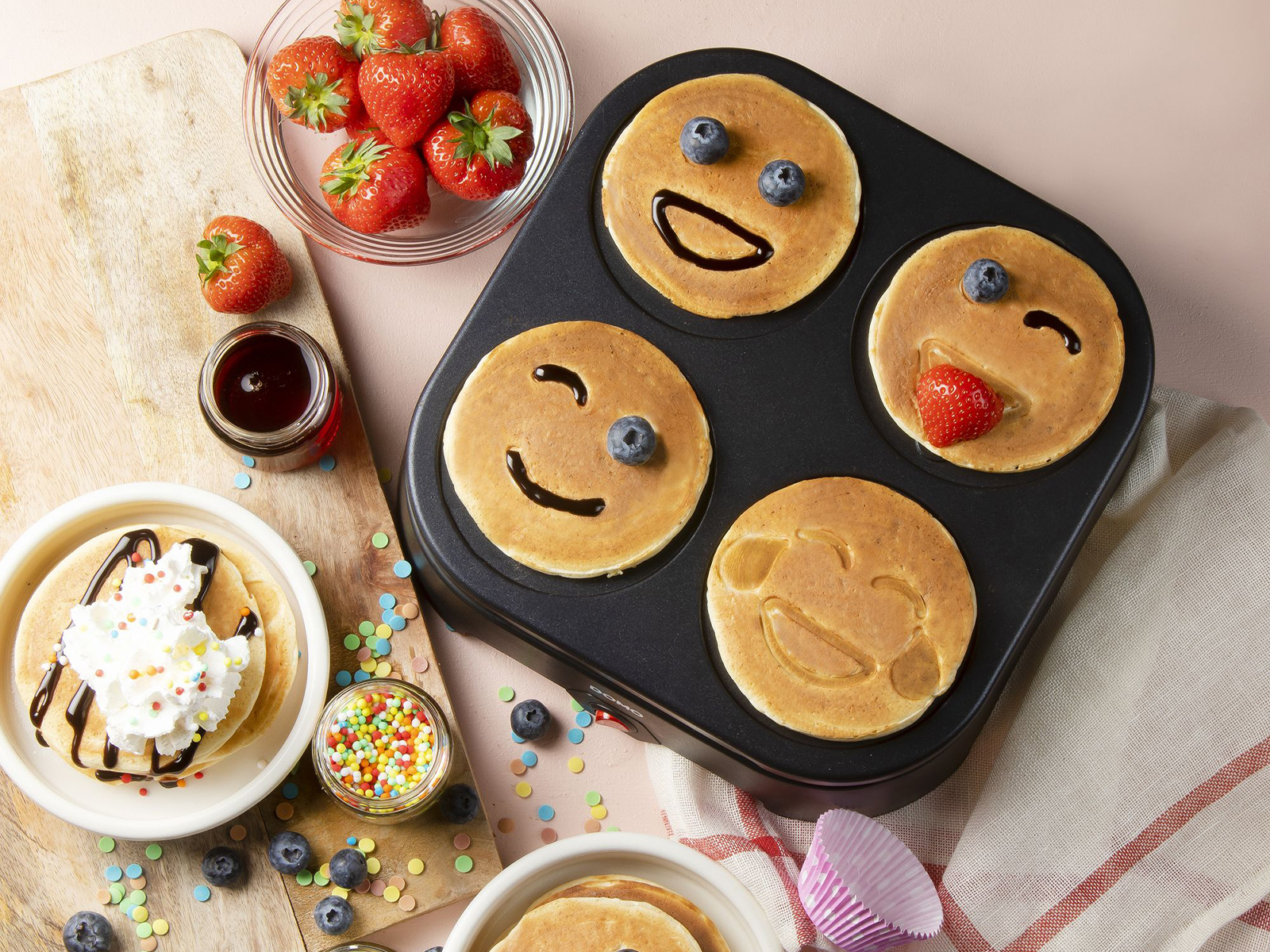 Naleśniki z uśmiechniętymi buźkami na patelni. Truskawki, syrop i posypka również są obecne. Różowe tło.