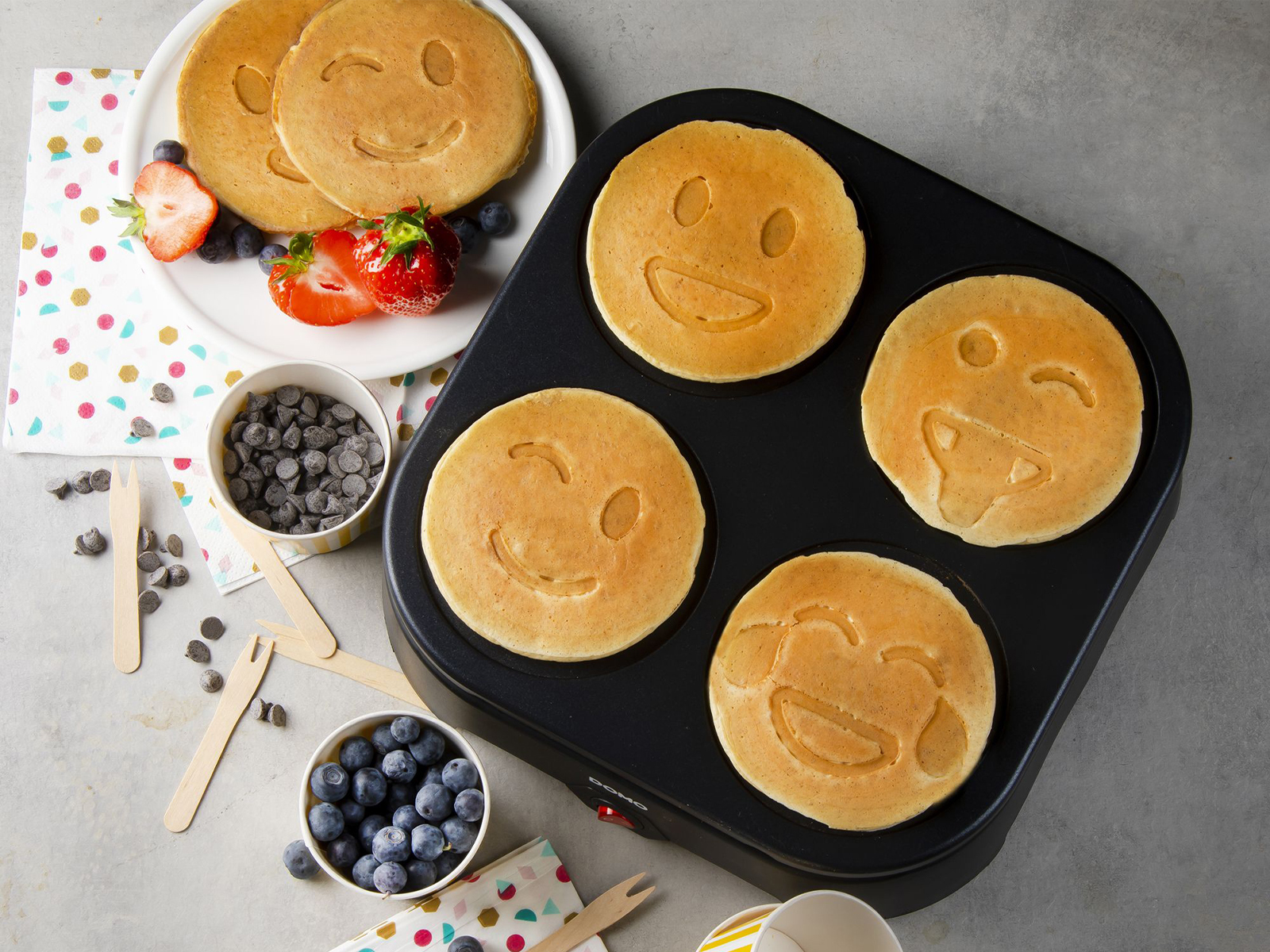 Maszyna do naleśników z naleśnikami ze smiley'ami, jagodami, kawałkami czekolady i truskawkami, na szarej powierzchni.