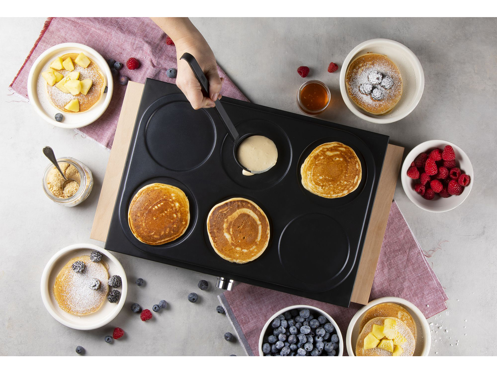 Pfannkuchen werden auf einer Grillplatte zubereitet. Verschiedene Beläge wie Beeren und Sirup sind vorhanden.