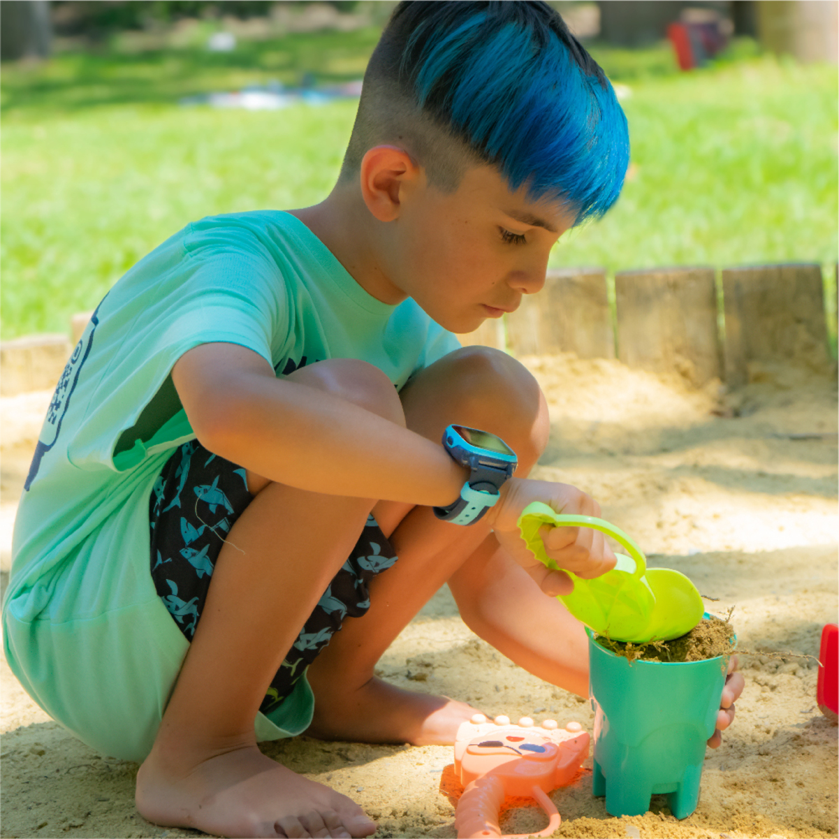 Un niño con cabello azul jugando en una caja de arena con un cubo y herramientas verdes.