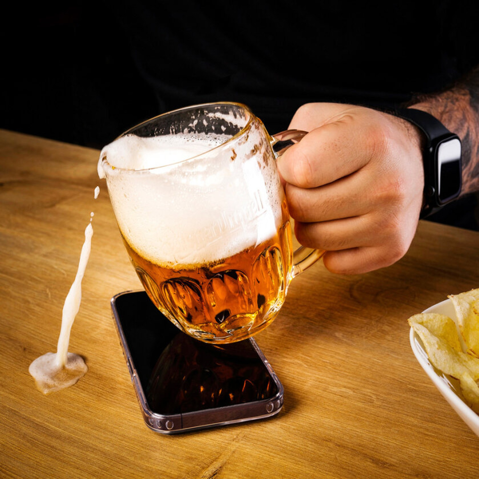 Person hält Bierkrug, Bier läuft auf Telefon. Chips in einer Schüssel im Hintergrund.