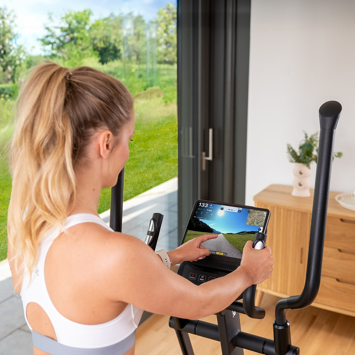 Frau trainiert auf einem Heimtrainer und schaut auf ein Tablet mit Fitness-App.