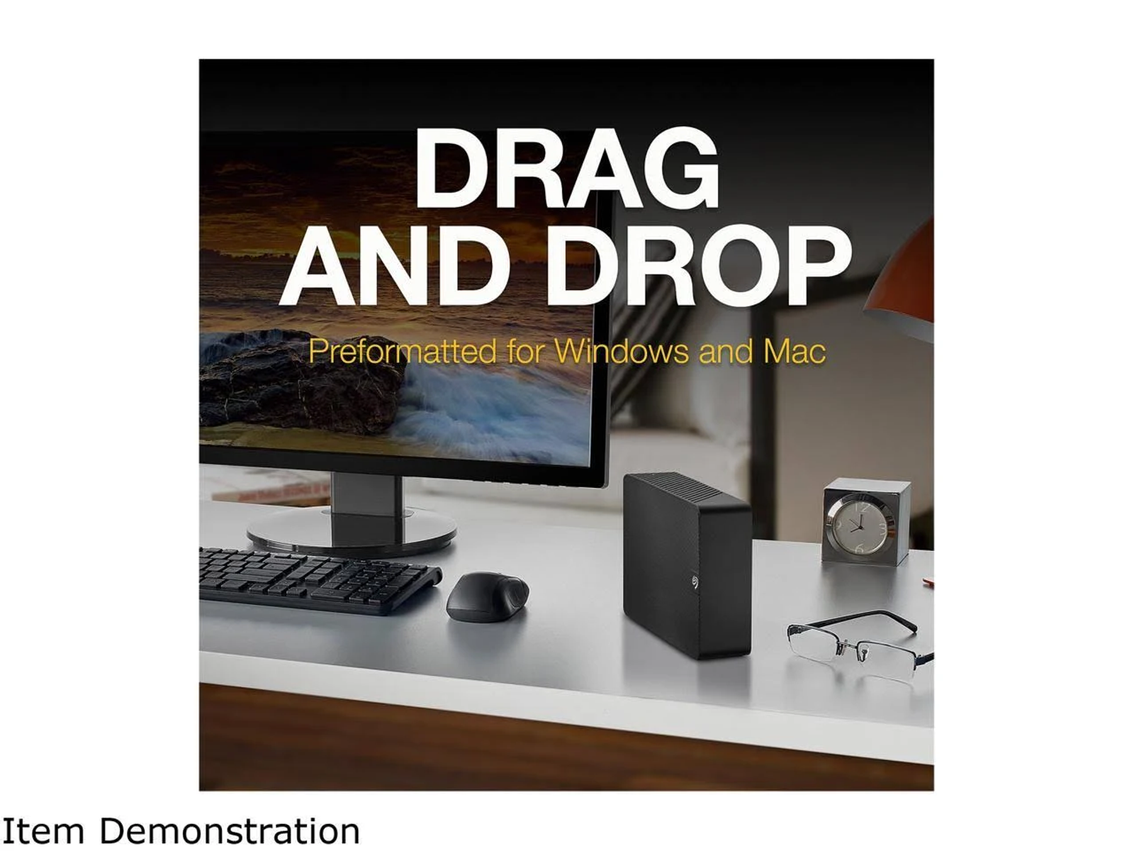 An external hard drive, mouse, keyboard, and glasses on a desk in front of a monitor.