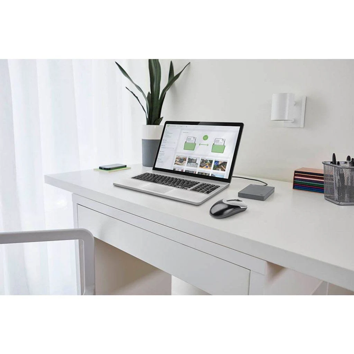 Laptop, mysz i hub na białym biurku z rośliną i oknem.