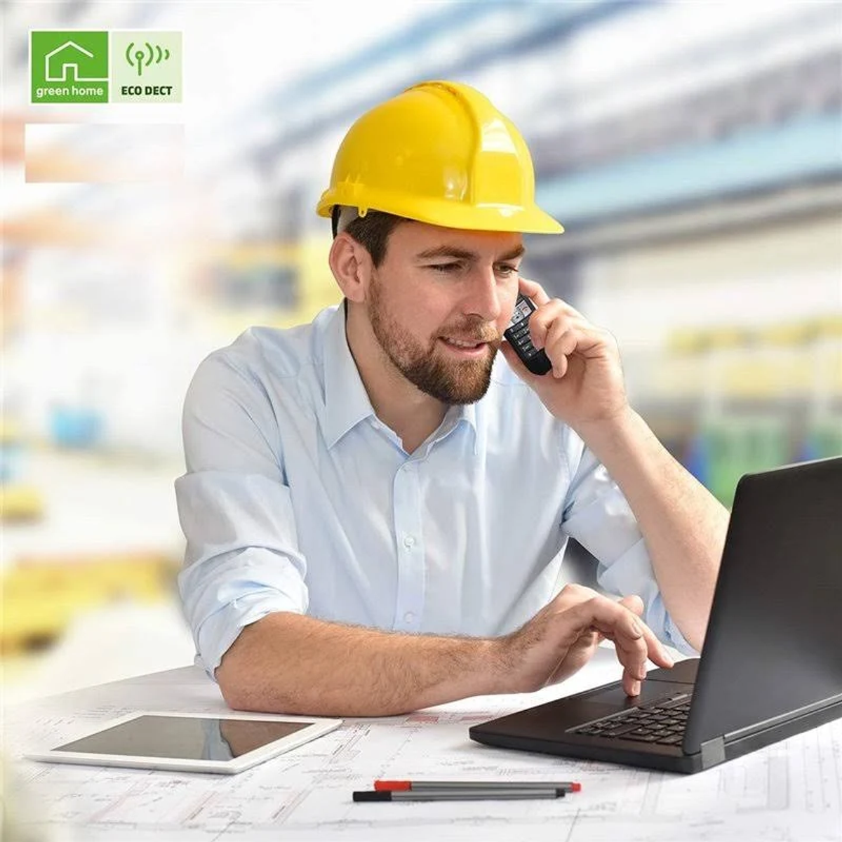 Mężczyzna w żółtym kasku rozmawia przez telefon i używa laptopa.