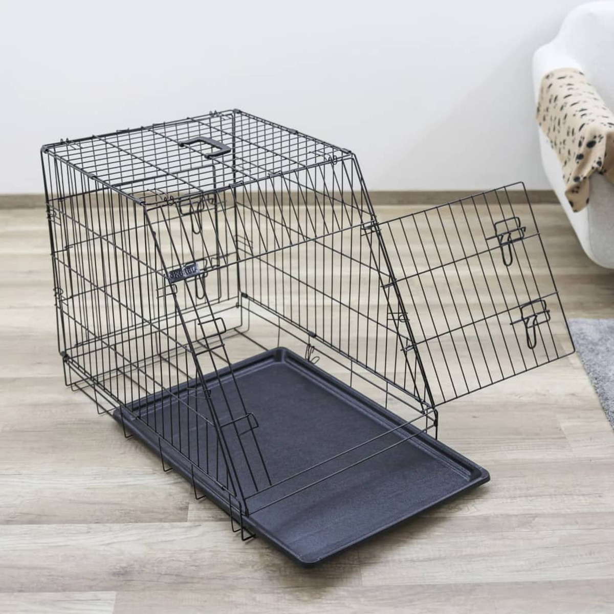 Eine offene schwarze Metall-Hundebox auf einem Holzboden. Im Hintergrund ein weißes Sofa und eine gemusterte Decke.