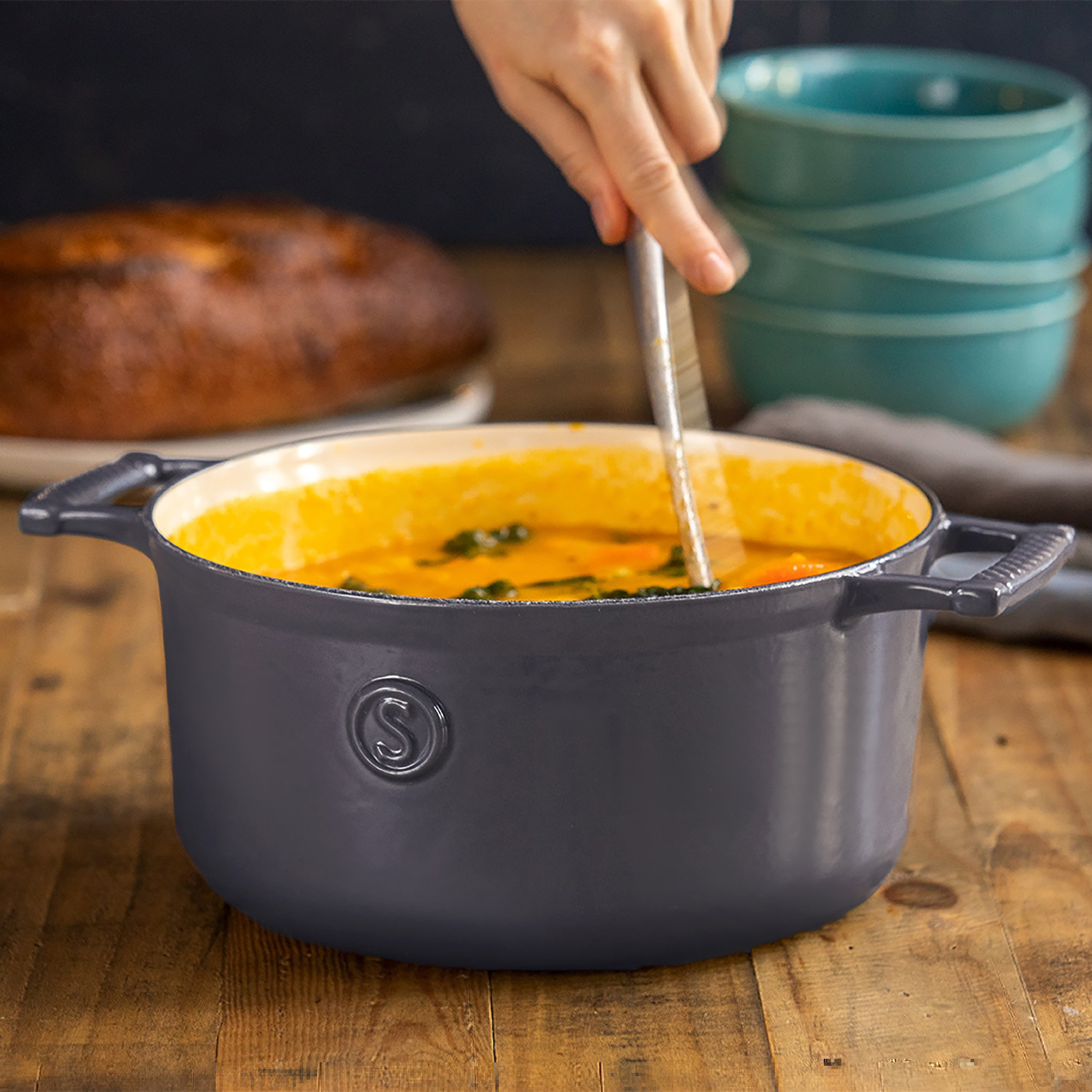 Eine Person rührt Suppe in einem grauen Topf auf einem Holztisch um. Brot und Schalen im Hintergrund.