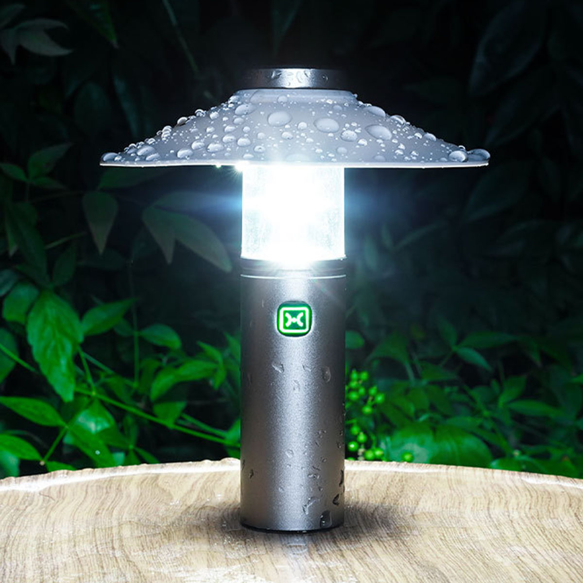 Eine silberne Lampe mit weißem Oberteil, beleuchtet. Es steht auf einer Holzoberfläche, im Hintergrund grüne Pflanzen.
